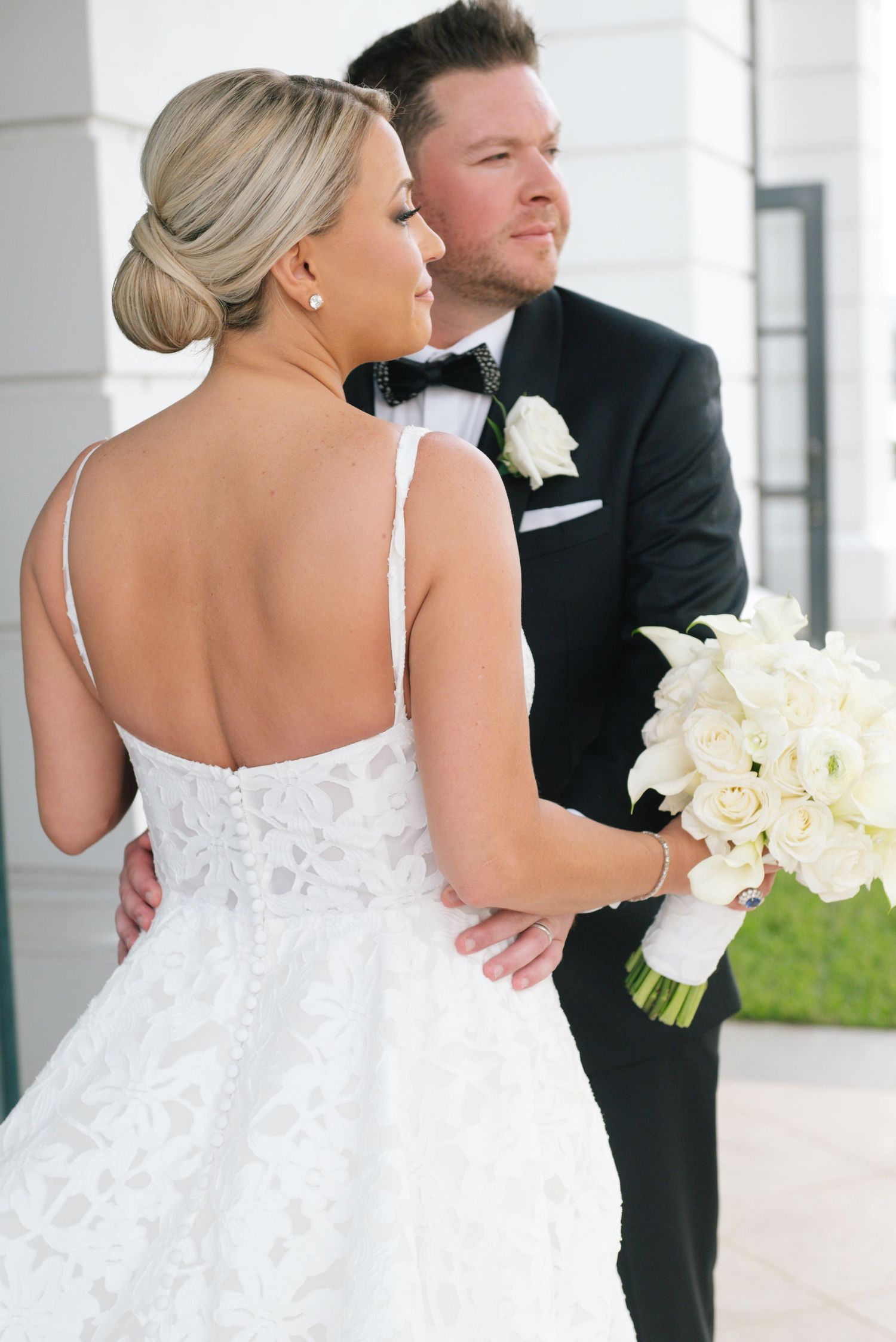 Bride in lace backless gown holds white bouquet as groom in tuxedo embraces her from behind.