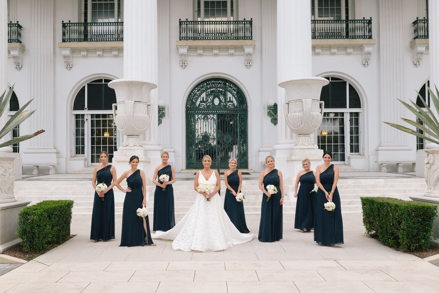 Bride in white gown with bridesmaids in navy dresses outside elegant white mansion.