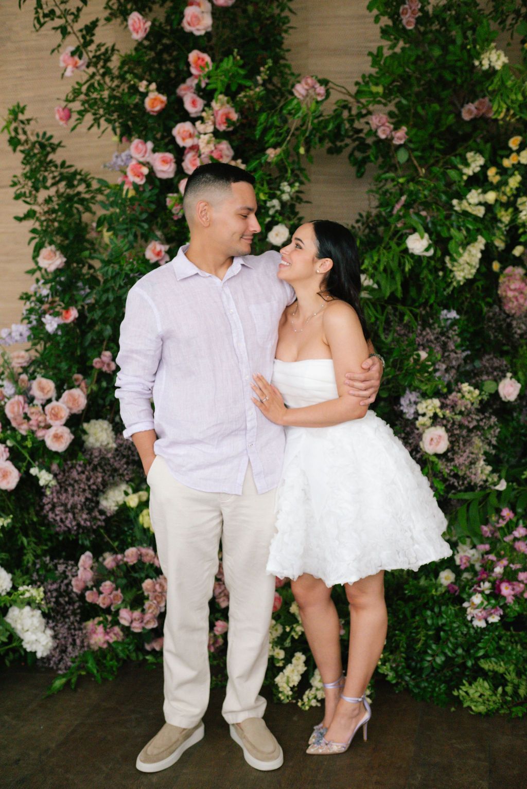 Couple posing against romantic floral wall with pink roses and greenery.
