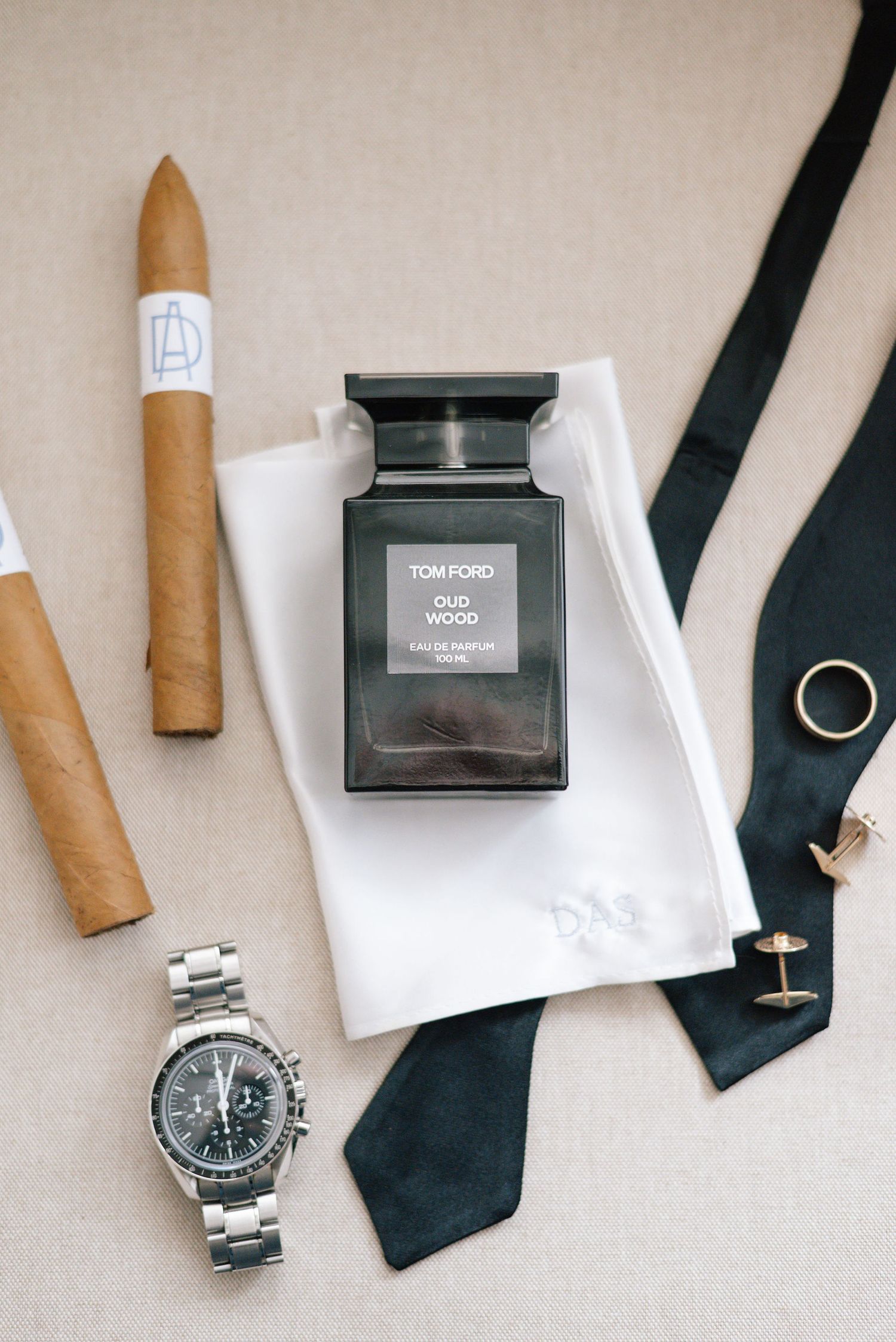 Elegant wedding details including a watch, black tie, cigars and Tom Ford cologne arranged on light background.
