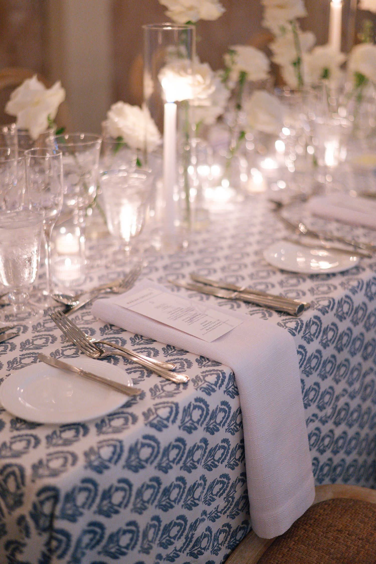 Elegant table setting with blue patterned tablecloth, white napkins, glassware and glowing candle centerpieces.