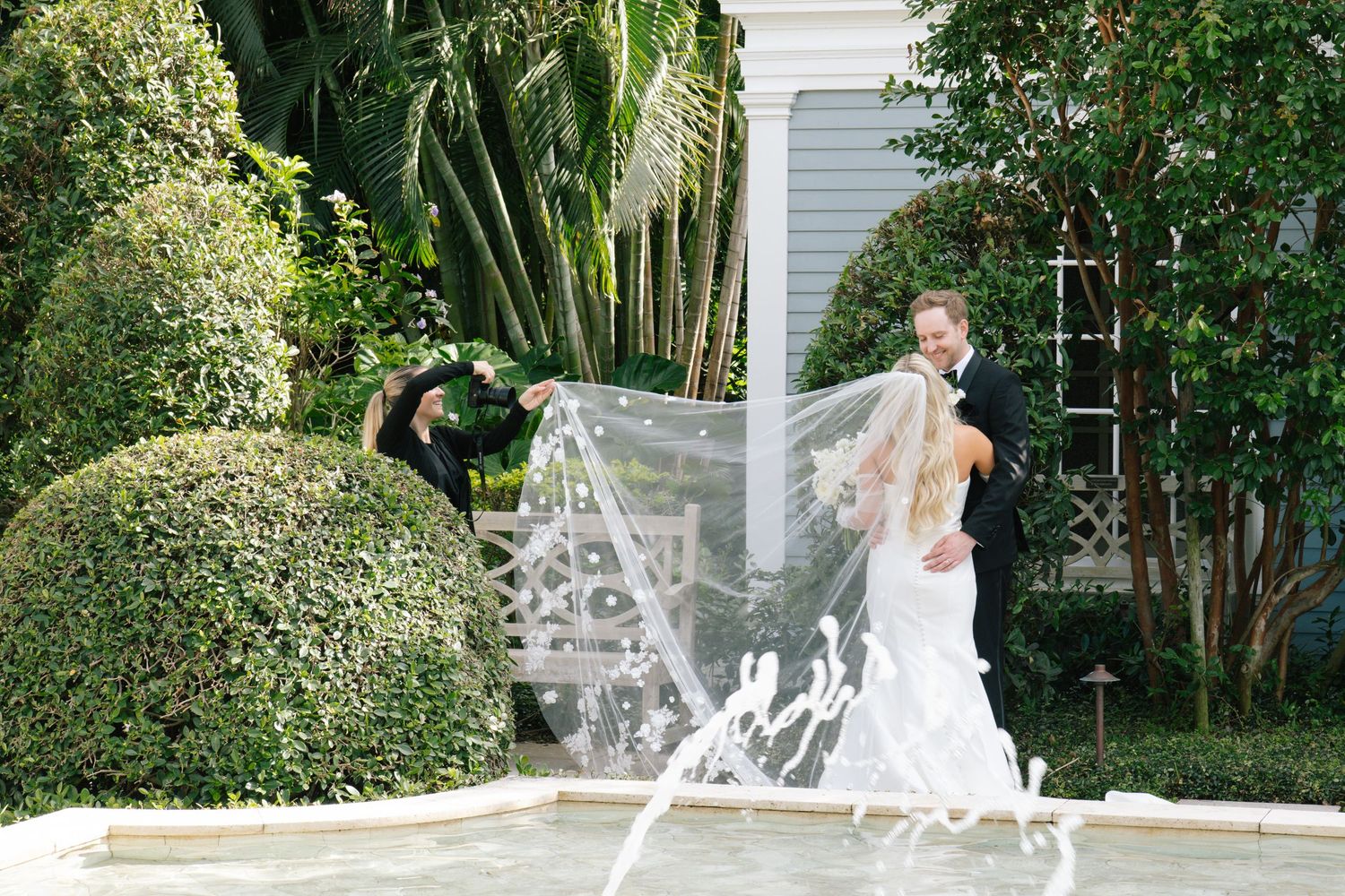 Wedding couple poses outdoors with flowing veil creating dramatic effect against garden backdrop.
