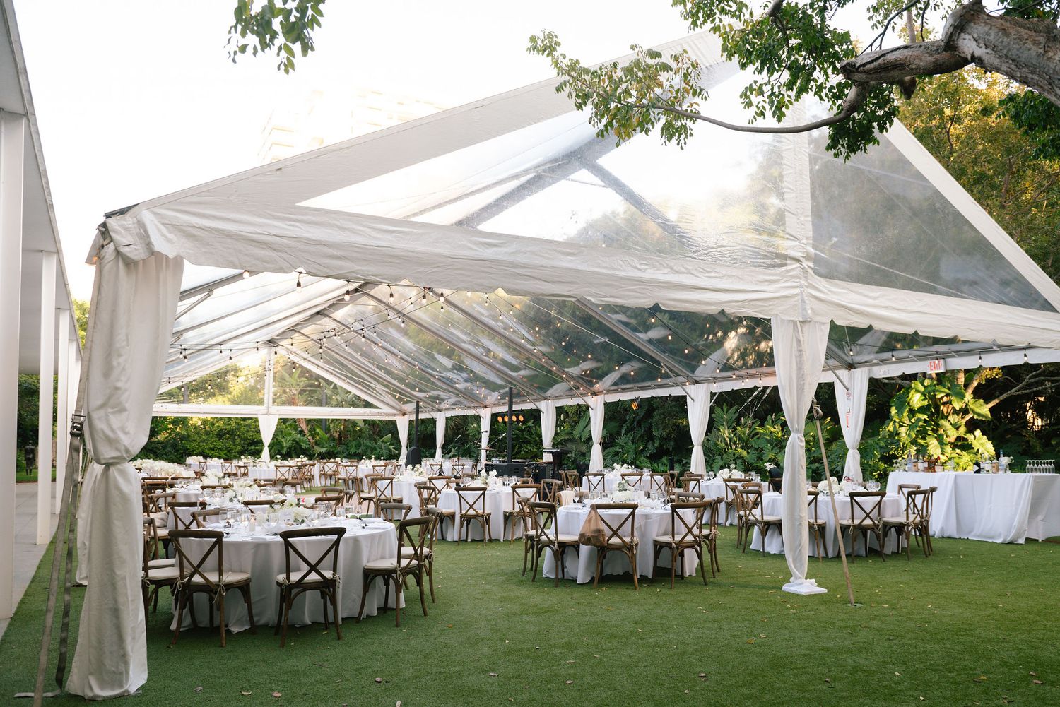 White wedding reception tent setup with elegant table arrangements and cross-back chairs on a manicured lawn.