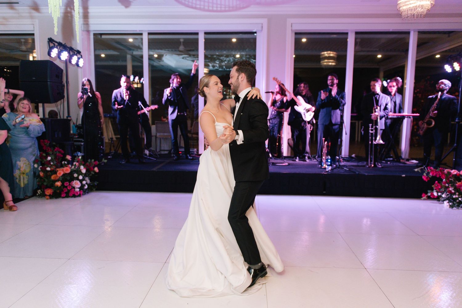 Couple sharing their first dance on illuminated white floor with band performing in background.