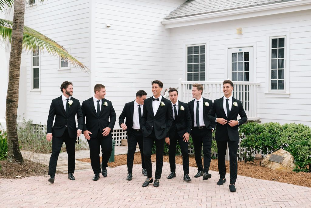 Wedding party members in black suits walk along white building exterior with palm trees.