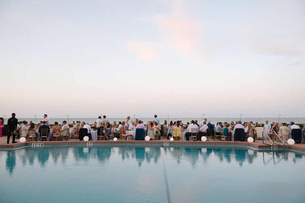 Panoramic view of wedding guests gathered by a pool during sunset at a coastal venue.
