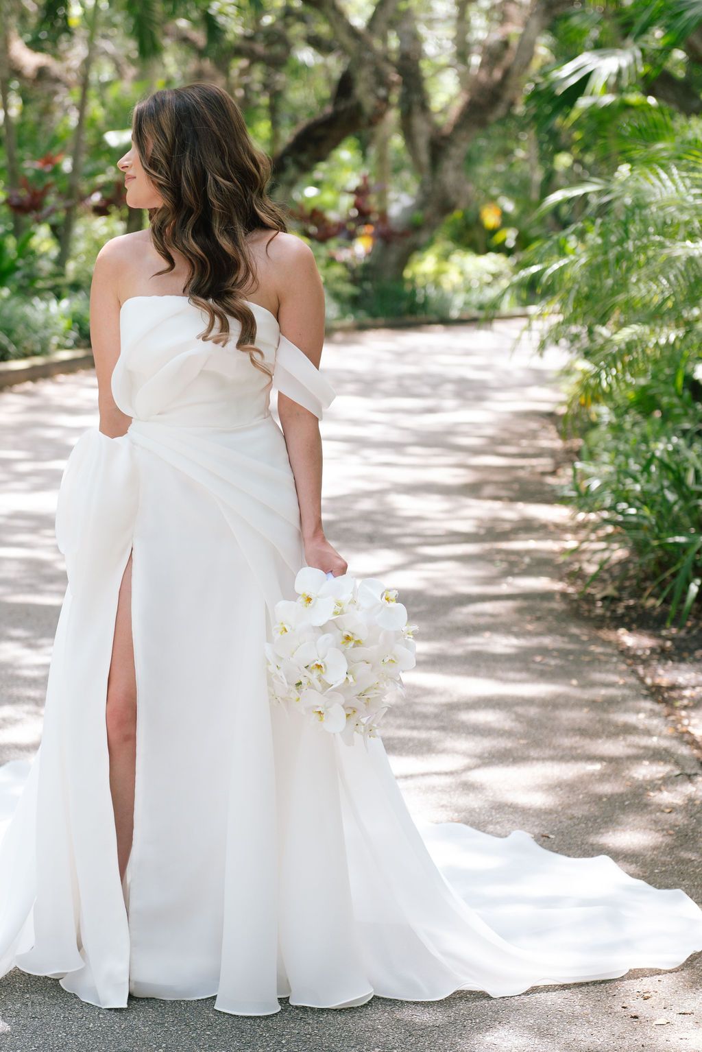 Elegant white wedding dress with flowing train photographed in a sunlit garden path.