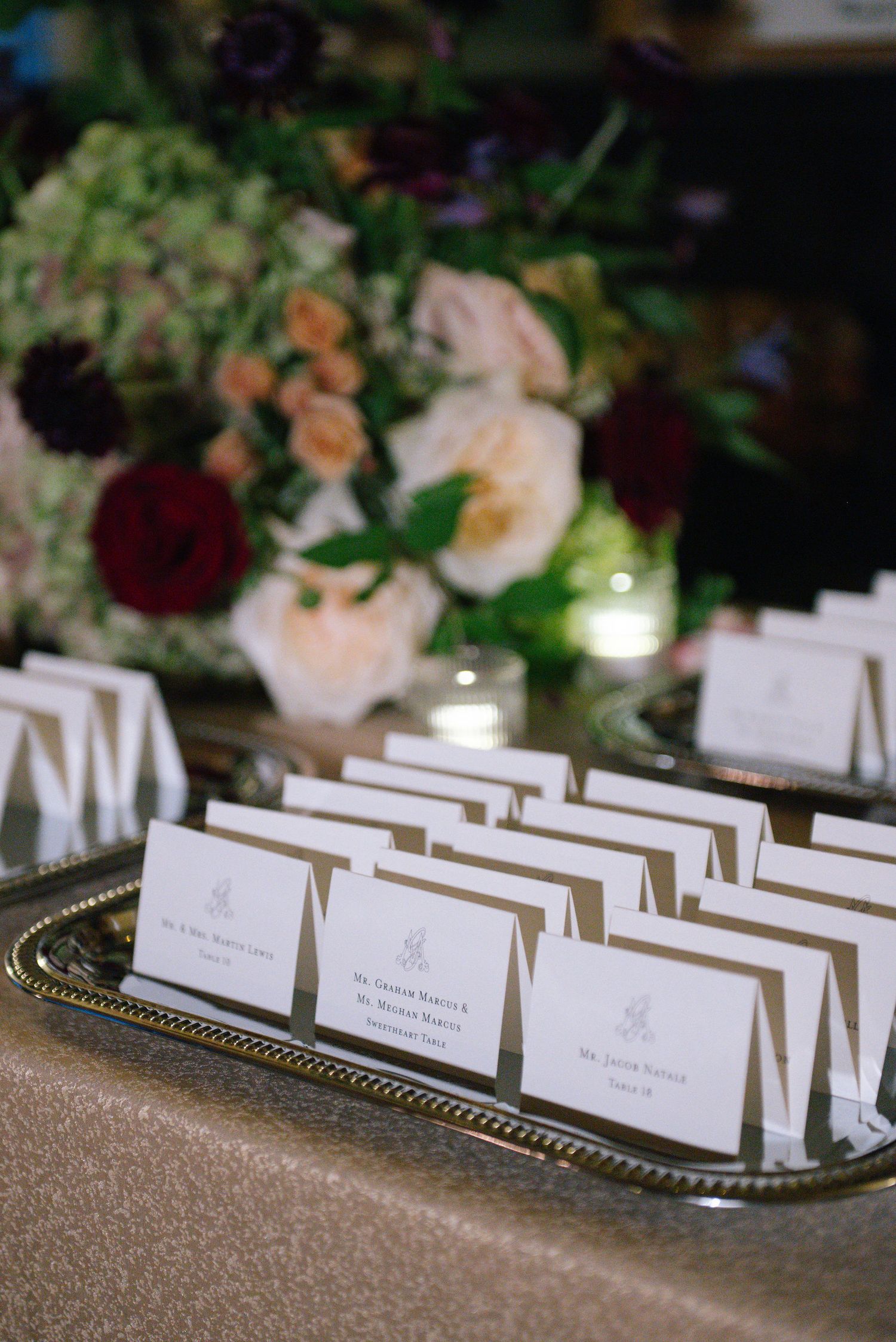 Wedding place cards arranged neatly on silver tray with floral arrangements in background.