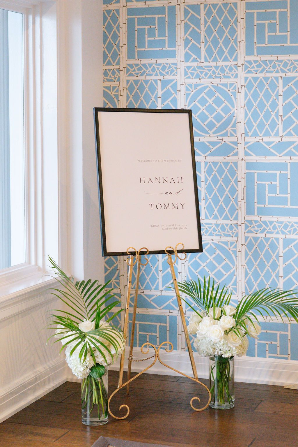 Elegant wedding signage with names displayed on stands against a blue geometric wallpaper with white floral arrangements.