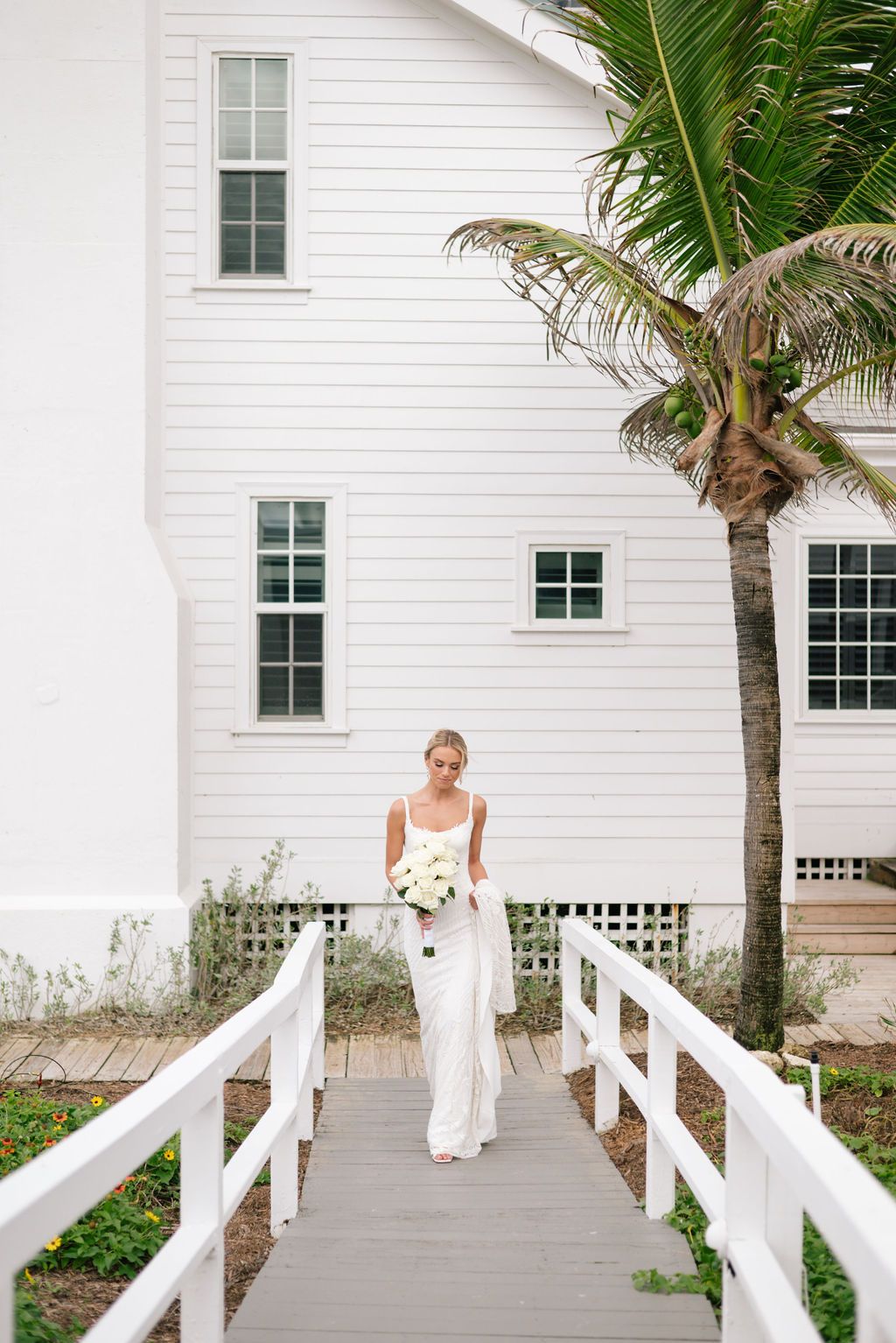 Bride walking along white wooden boardwalk with palm trees and coastal architecture.