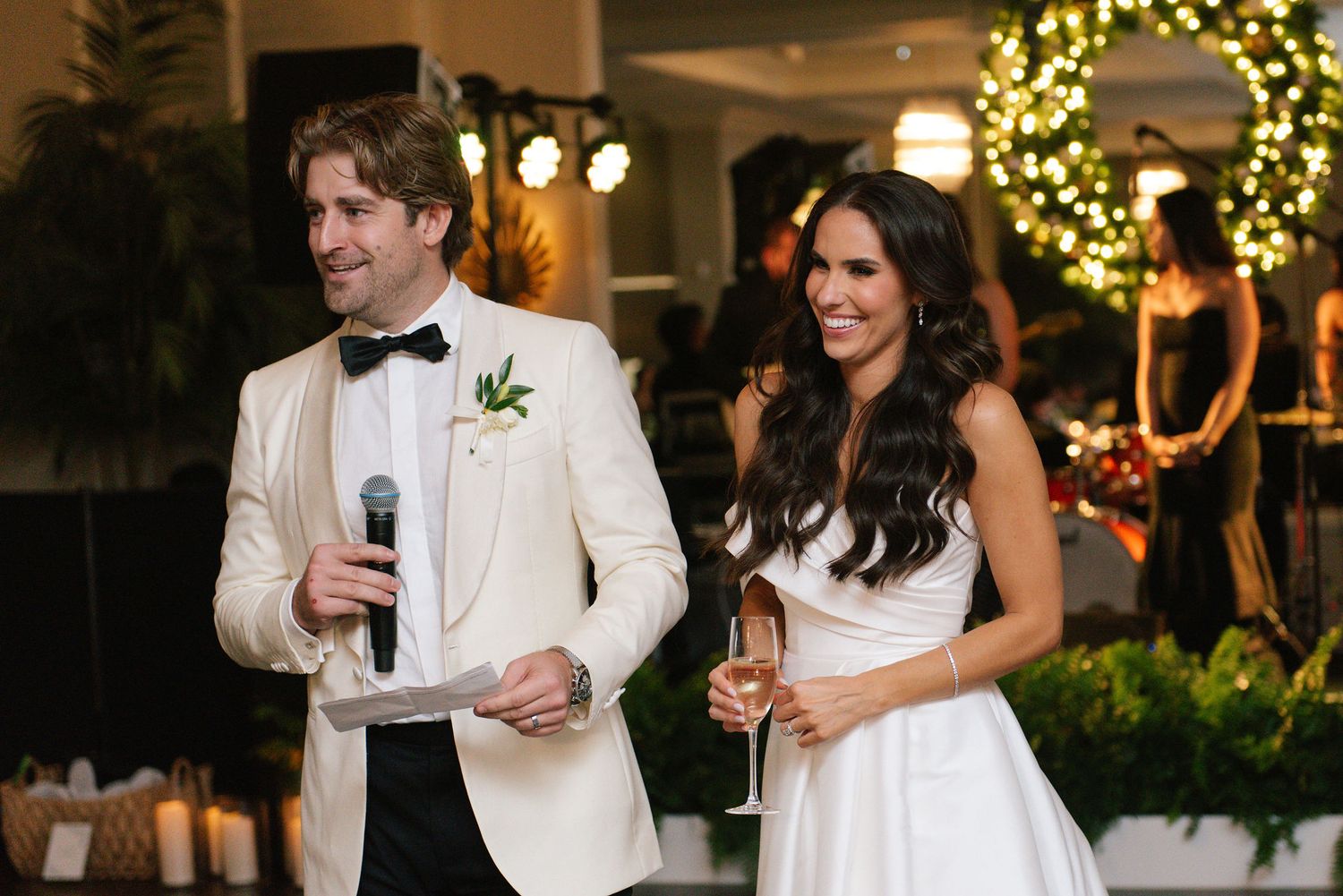 Newlyweds giving a speech at their wedding reception with holiday decor in background.