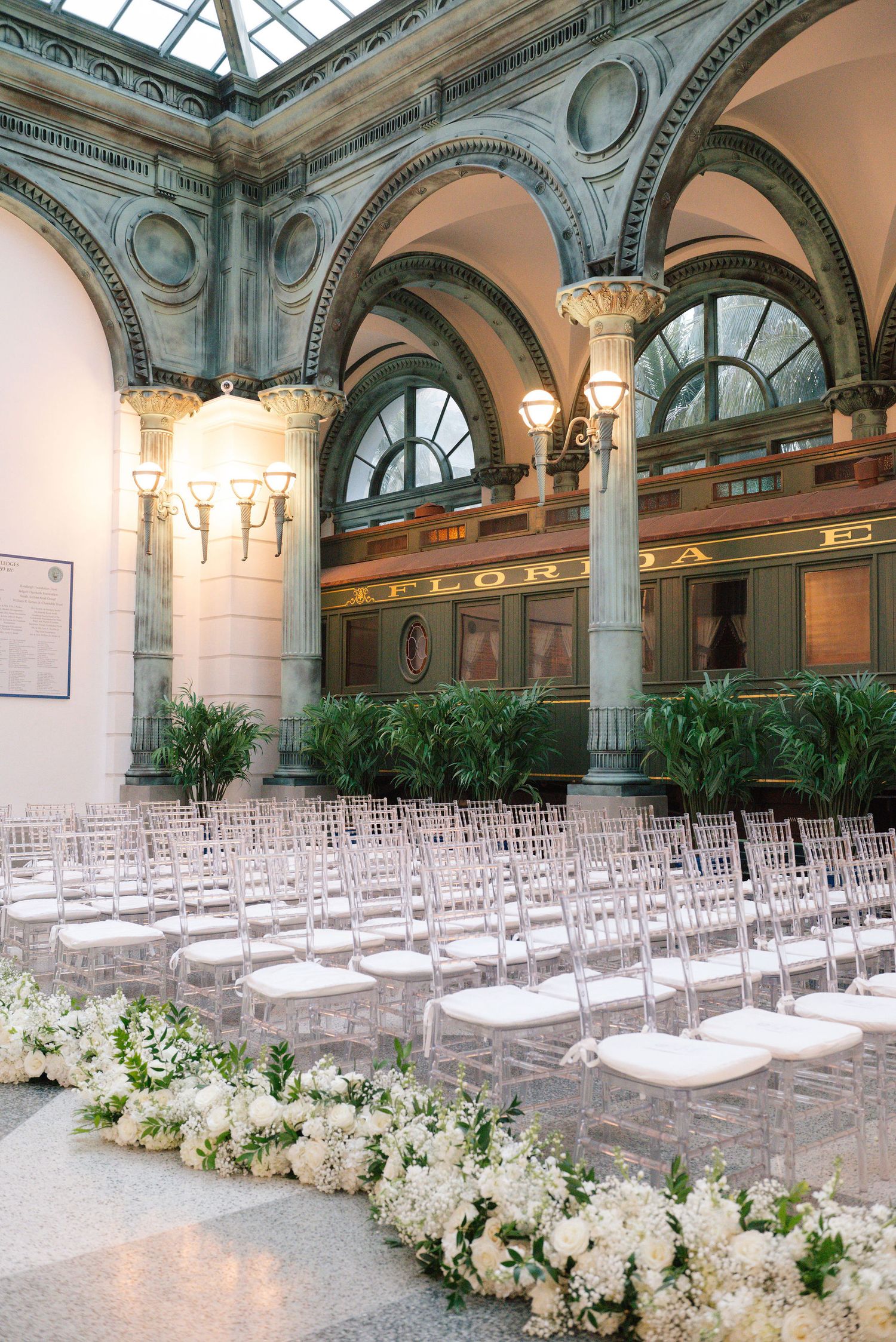 Classic ballroom wedding setup with ghost chairs and lush white floral aisle decorations.