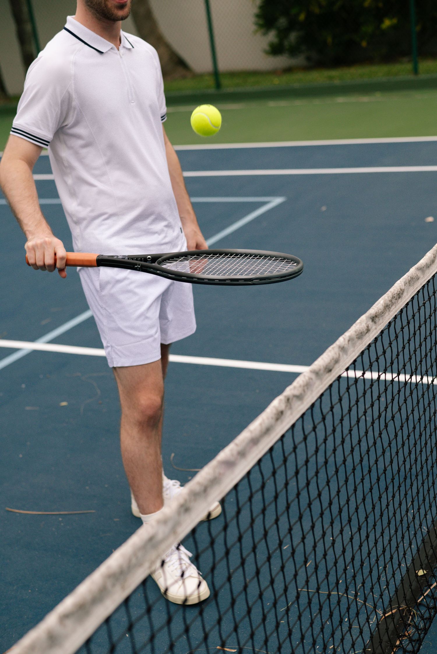 Tennis player in white outfit serves ball on outdoor blue court with net.