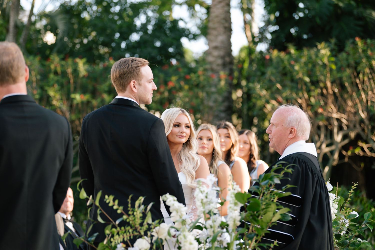 Wedding ceremony in progress under palm trees with officiant and wedding party at tropical venue.
