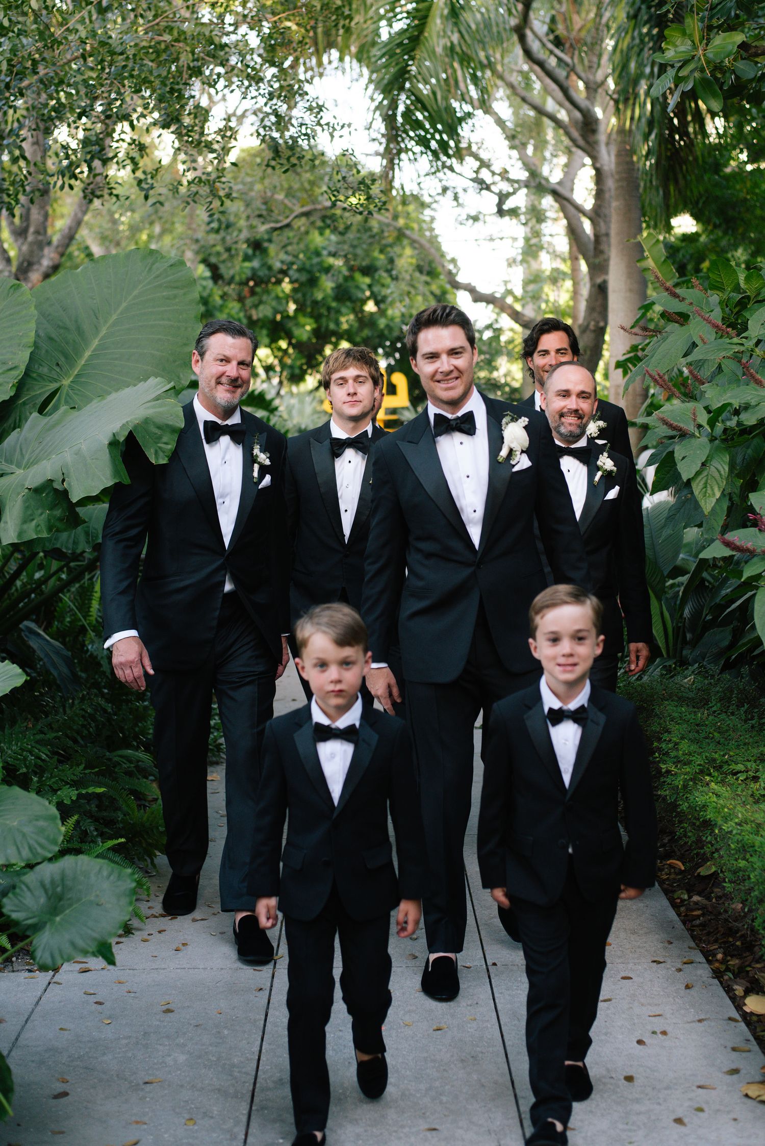Groomsmen in black tuxedos walk together down a garden path lined with lush greenery.