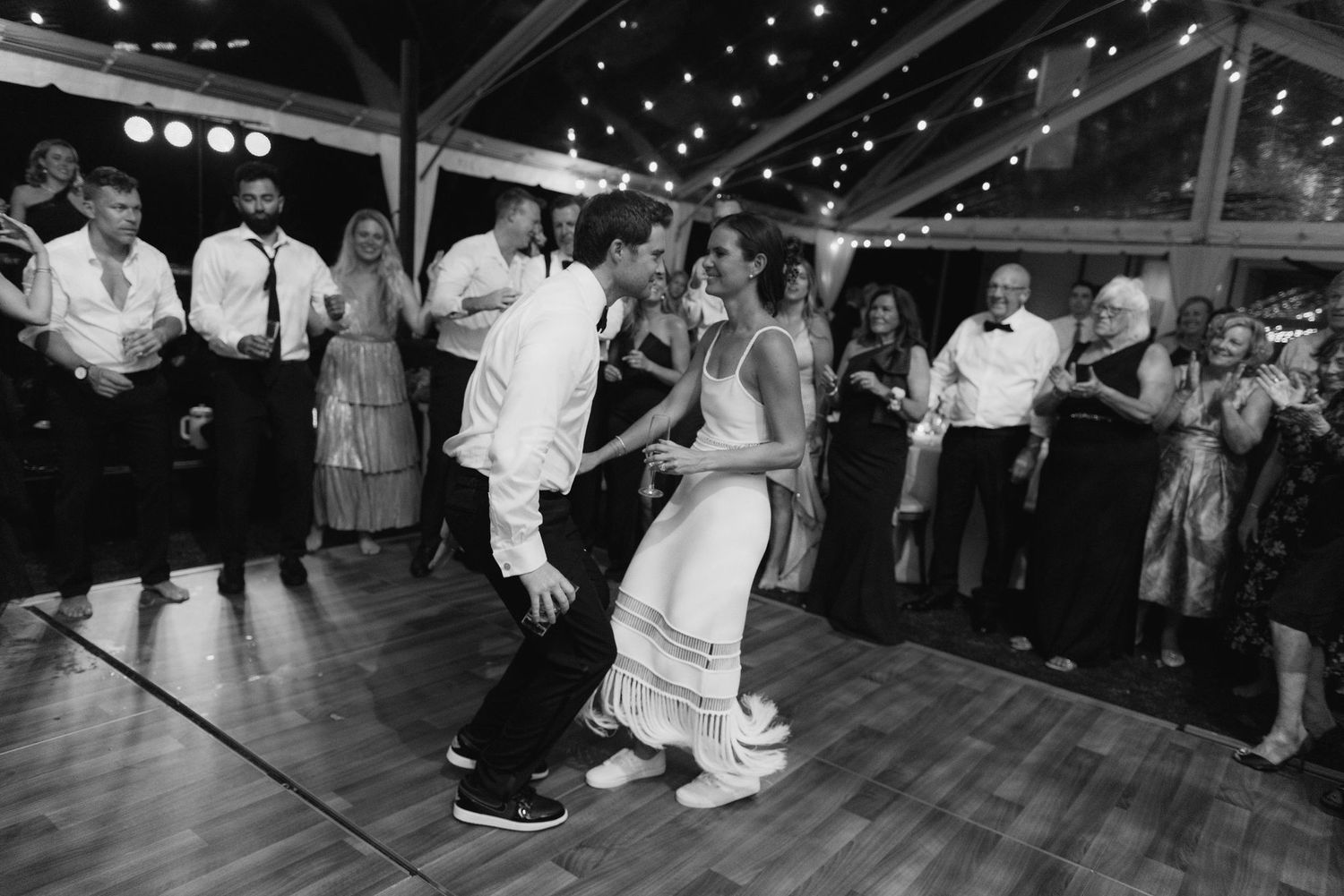 Wedding guests dance and celebrate on a dark dance floor under string lights.