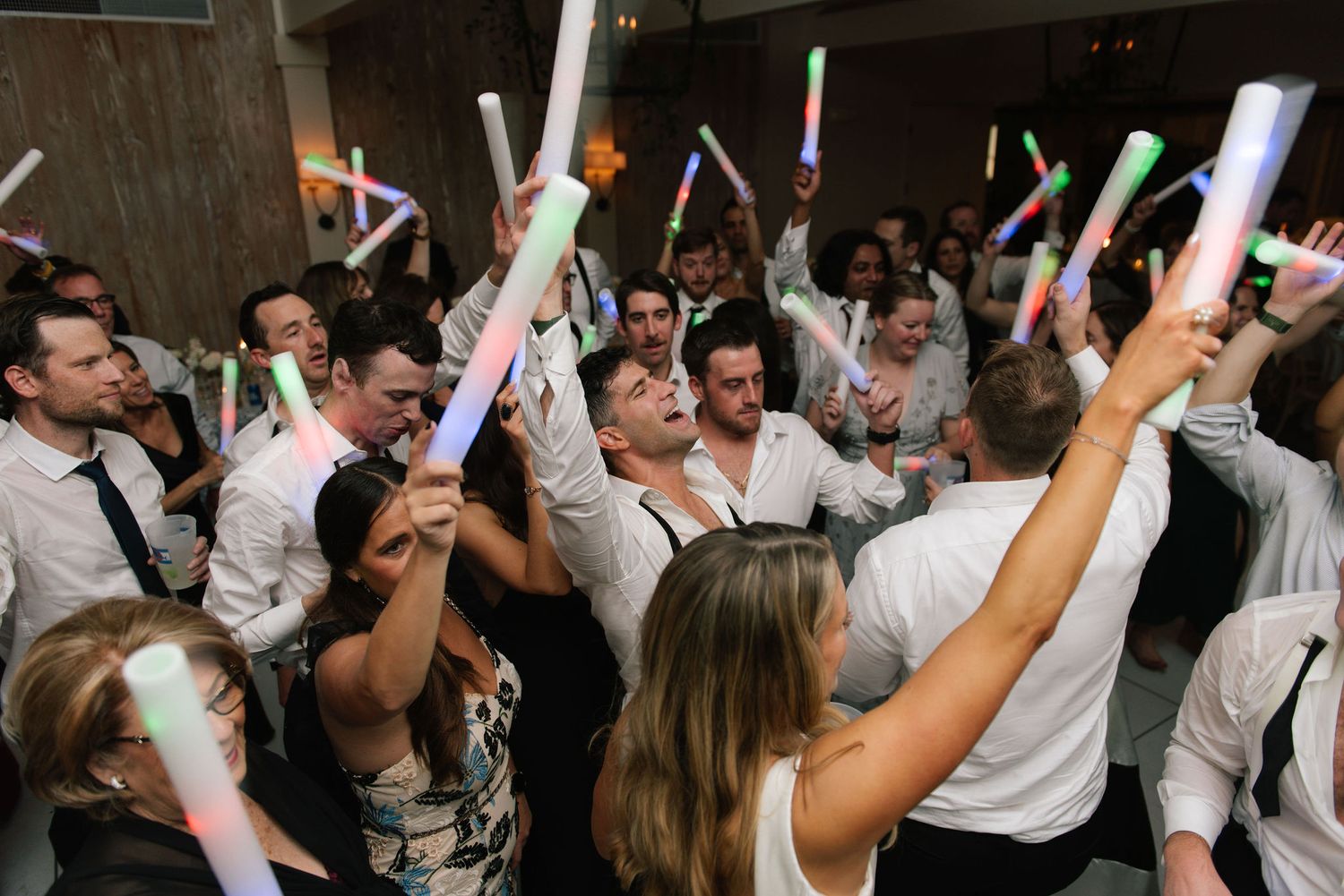 Crowd of wedding guests dancing and celebrating with glowing light sticks on the dance floor at a reception party.