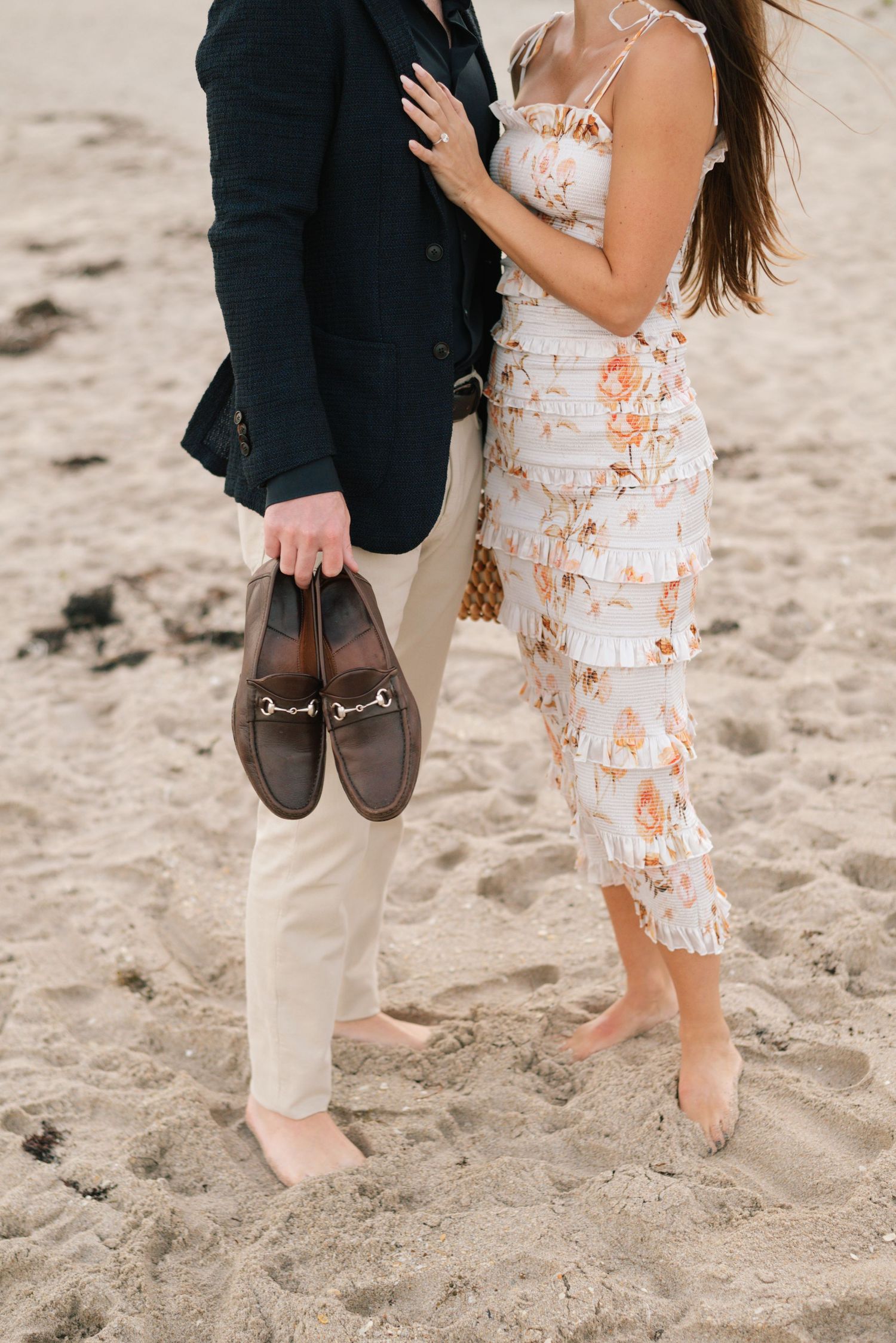 Barefoot couple walking on beach sand holding dress shoes, wearing formal and casual attire.