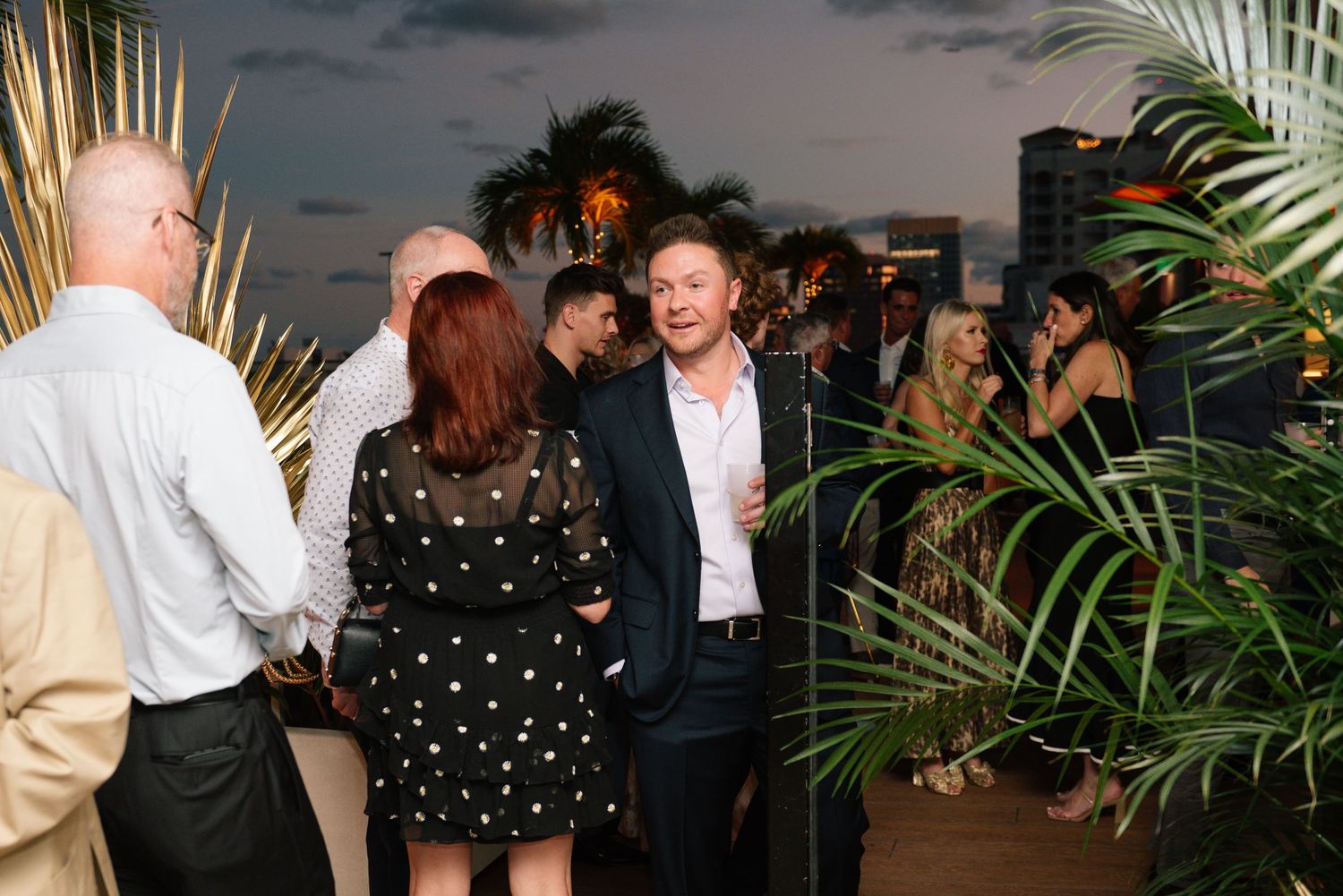 Evening party scene with guests mingling among tropical palm decorations.