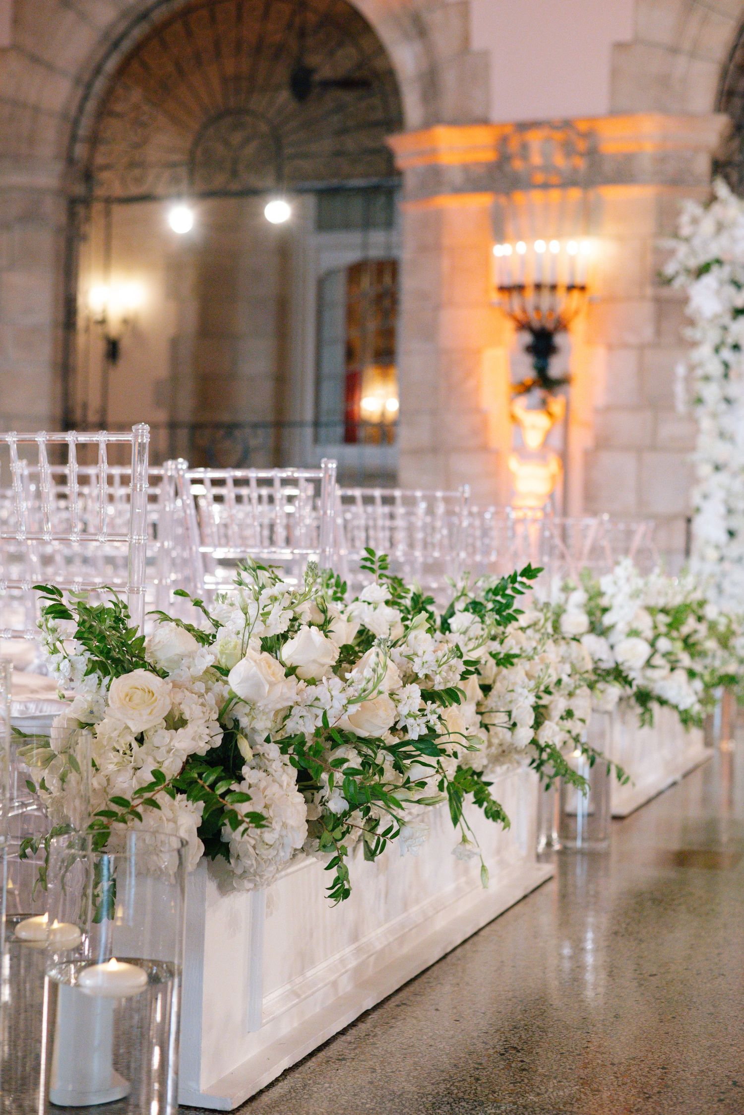 Lush white floral arrangements with greenery line wedding ceremony aisle with candles.