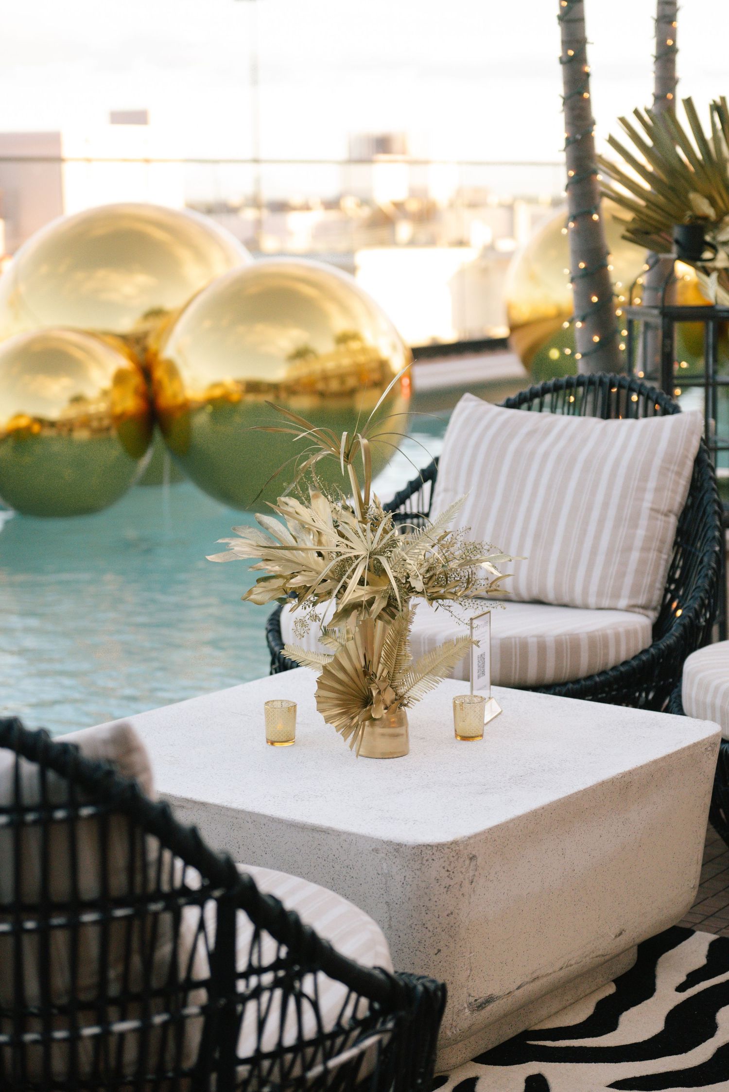 Poolside seating area with white cushions and dried palm arrangements against reflective gold spheres.