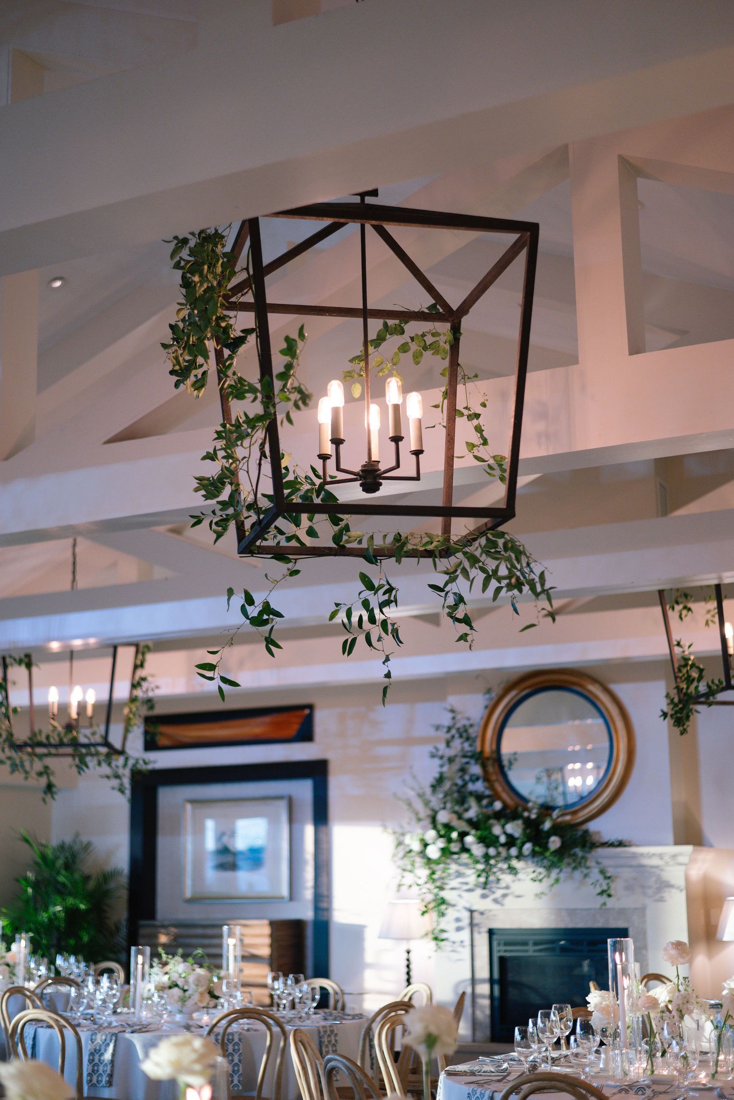 Elegant wedding reception venue with lantern chandelier and greenery vines decorating white beamed ceiling.
