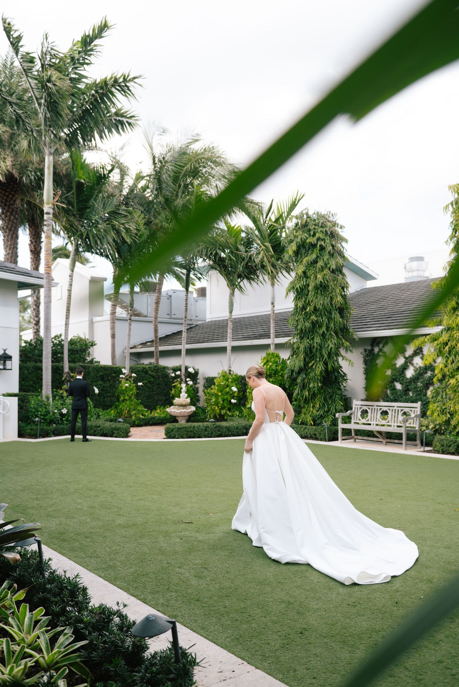 A white wedding dress flows elegantly across a lush green lawn with palm trees in the background.