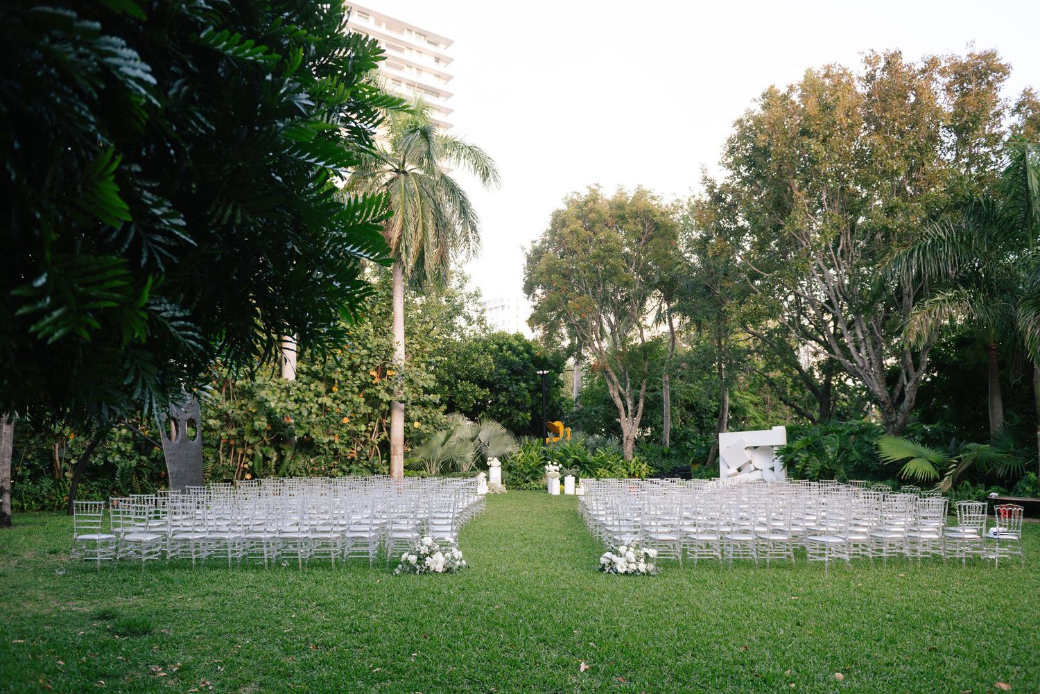 White chairs arranged in rows on a lush green lawn for an outdoor wedding ceremony with palm trees in the background.