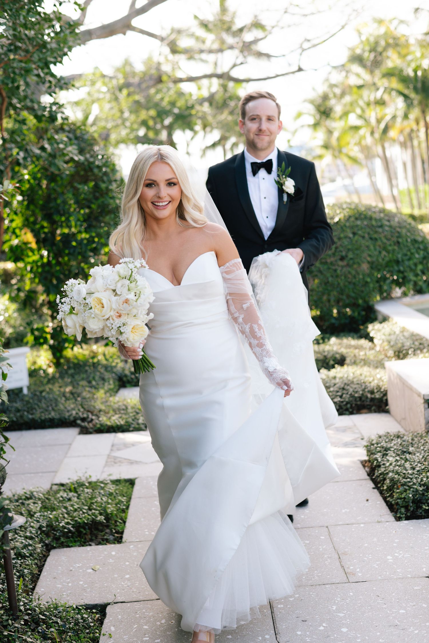 Bride in flowing white gown twirls on a garden path while groom watches.