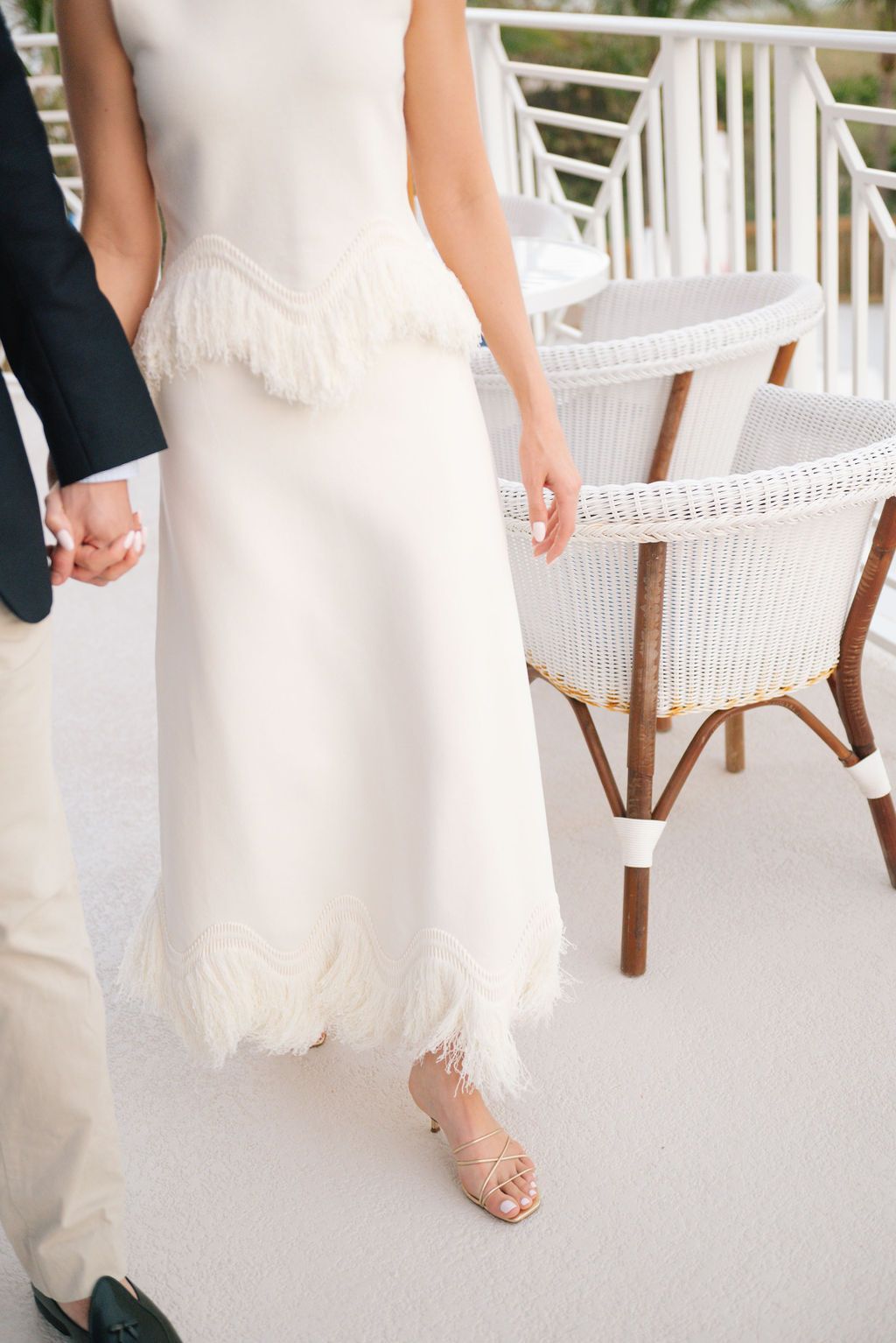 Close-up of elegant white dress with fringe detail and clasped hands on a coastal balcony.
