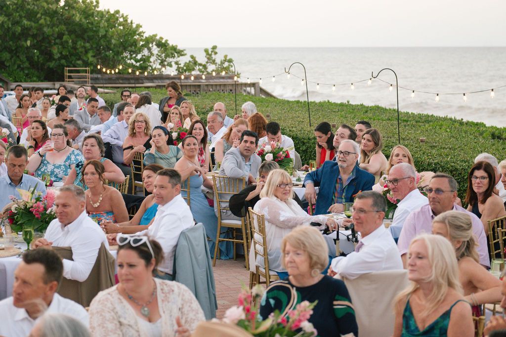 Wedding guests seated outdoors at round tables during a coastal reception.