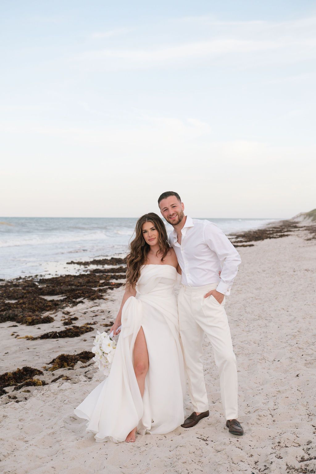 Romantic beach wedding photoshoot with couple in all white against ocean backdrop.