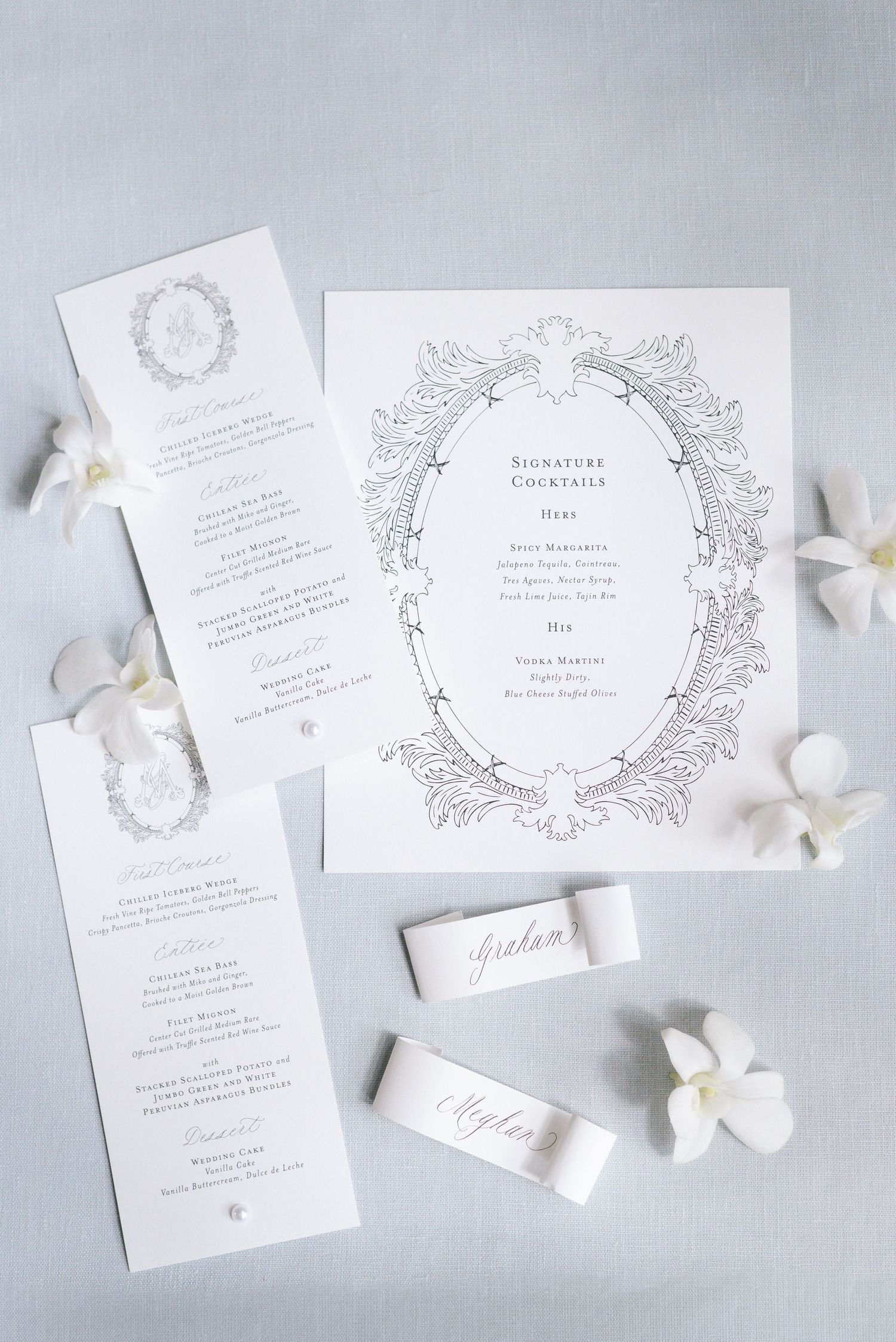 Ornate wedding invitation suite with floral wreath design and white petals on gray background.