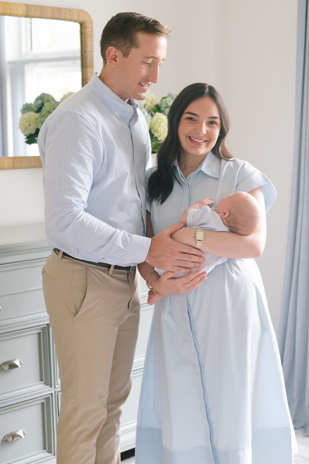 A couple in coordinating light outfits lovingly hold their newborn baby in a bright nursery setting.