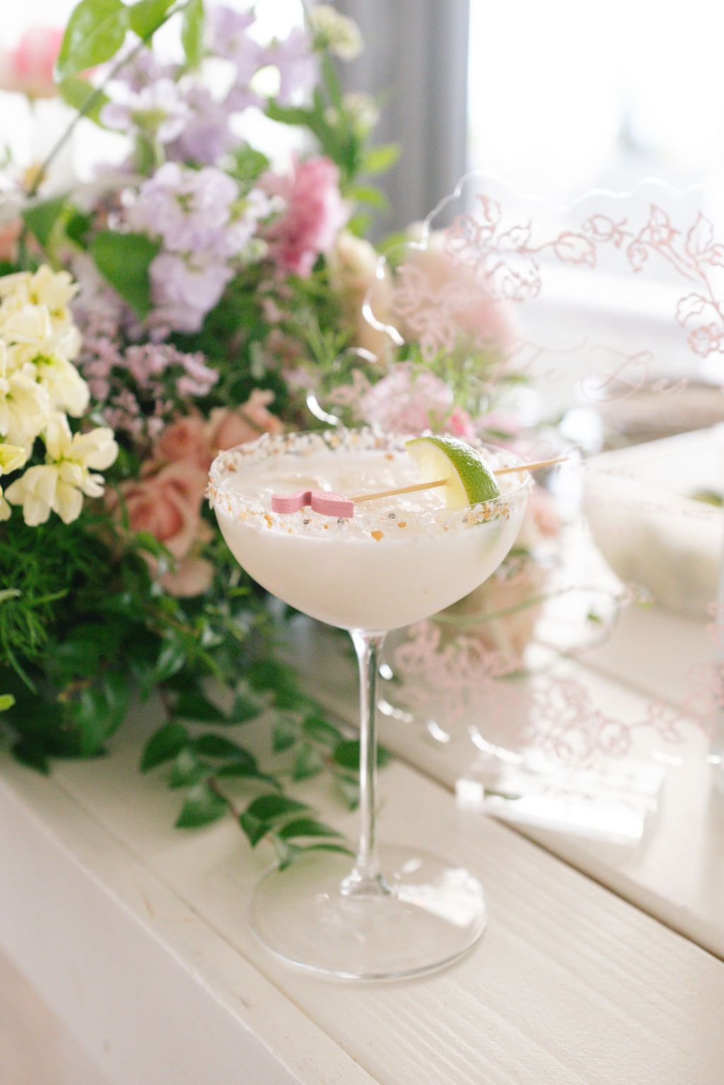 Elegant cocktail garnished with pink petals next to pastel floral arrangements on white table.