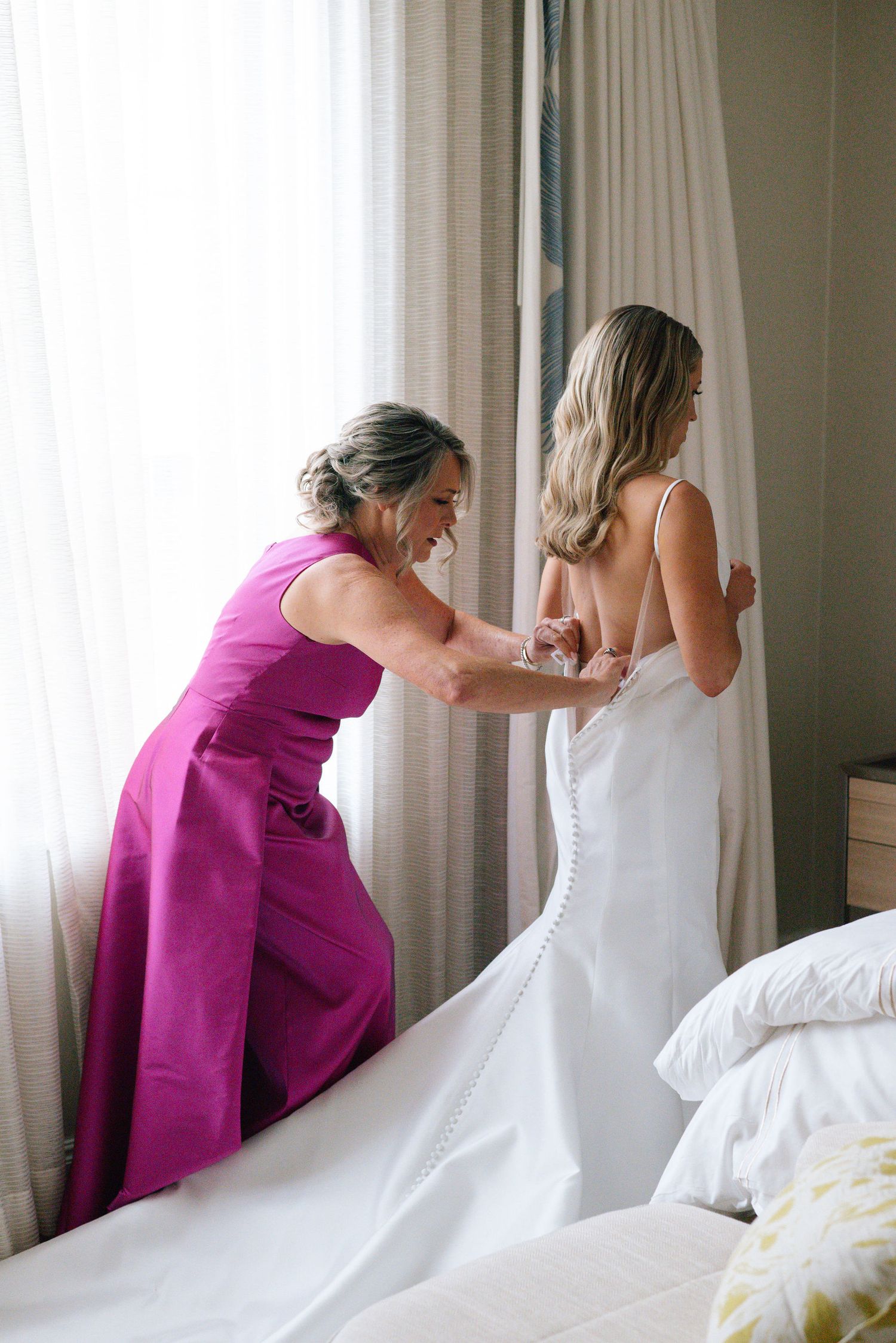 Series of photos showing a wedding dress being laced up with help from someone in a magenta dress.