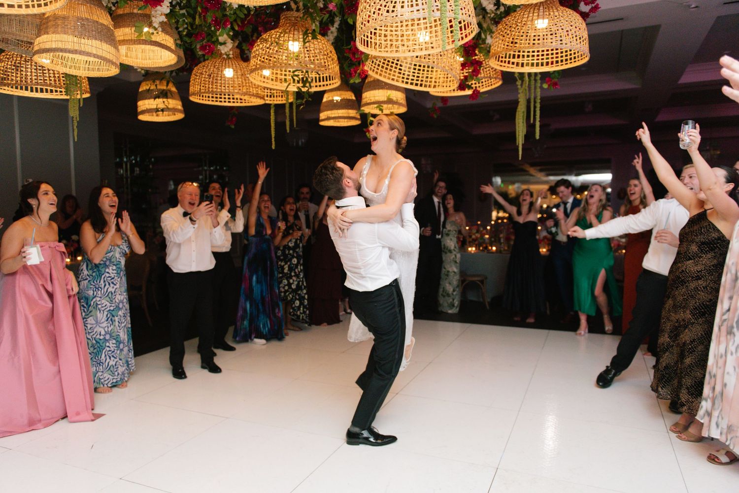 Wedding guests dancing on white dance floor under woven pendant lights.