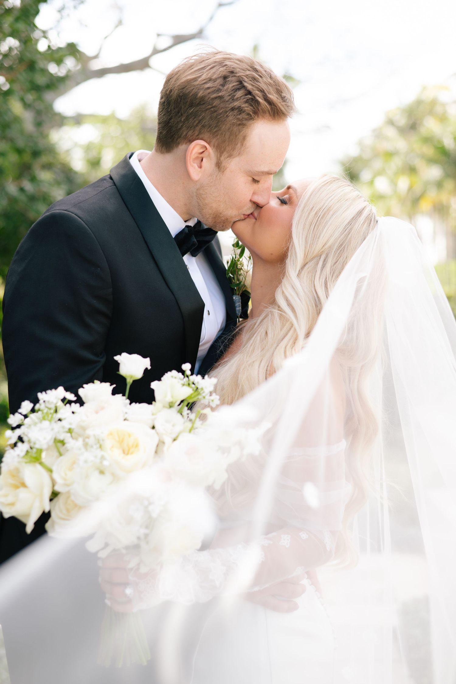 A romantic wedding couple sharing intimate kisses in formal attire with a white floral bouquet and veil.