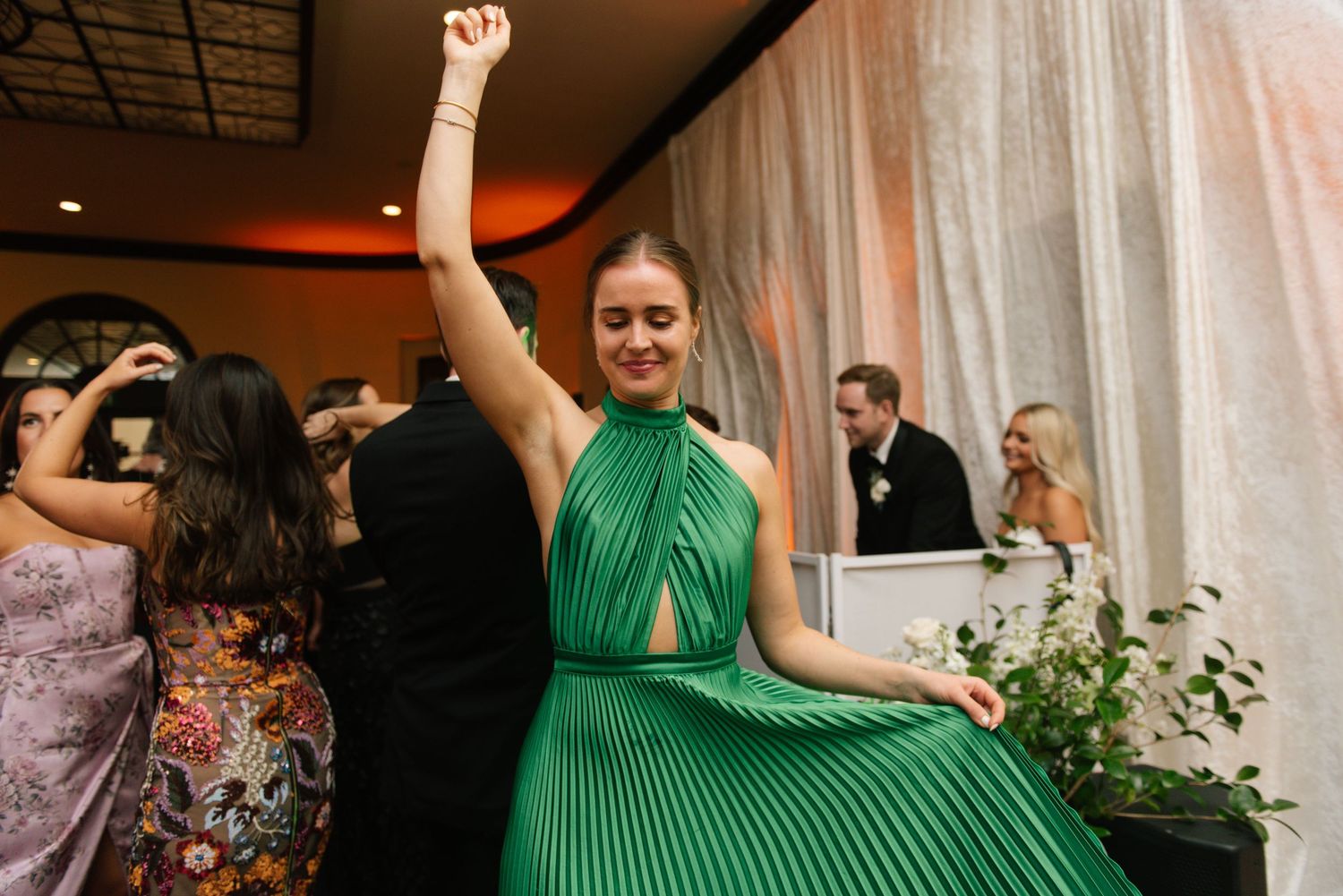 Woman in emerald green pleated halter dress dances joyfully at an indoor wedding reception.