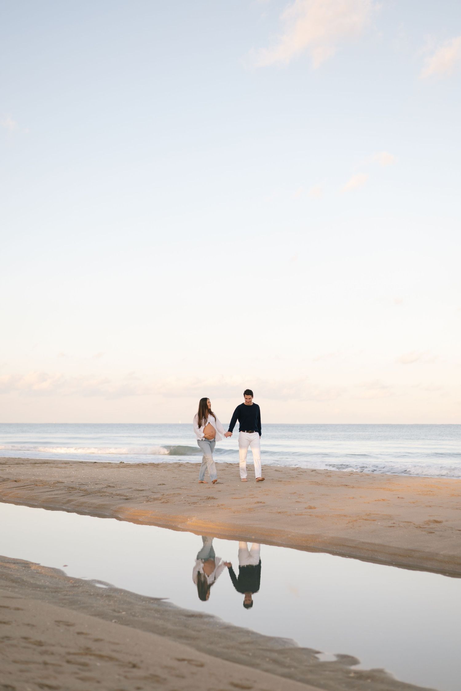 Couple walking along a beach at sunset with reflections in the wet sand.