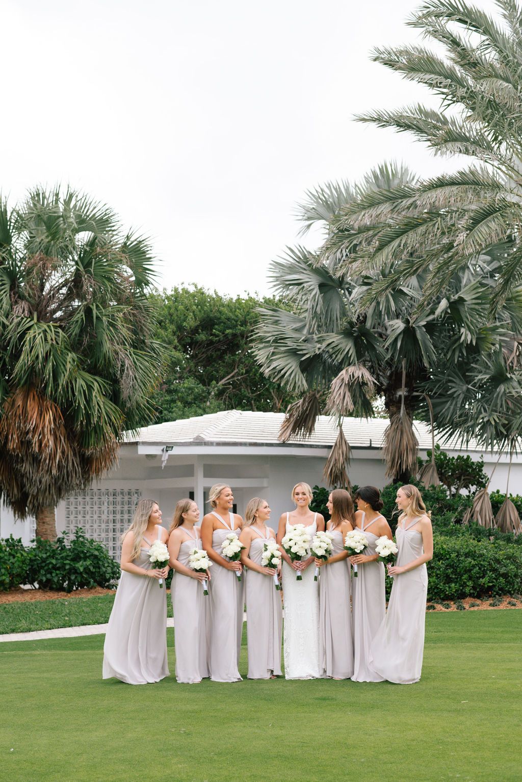 Bridesmaids in light gray dresses pose on a green lawn with palm trees and white building in the background.