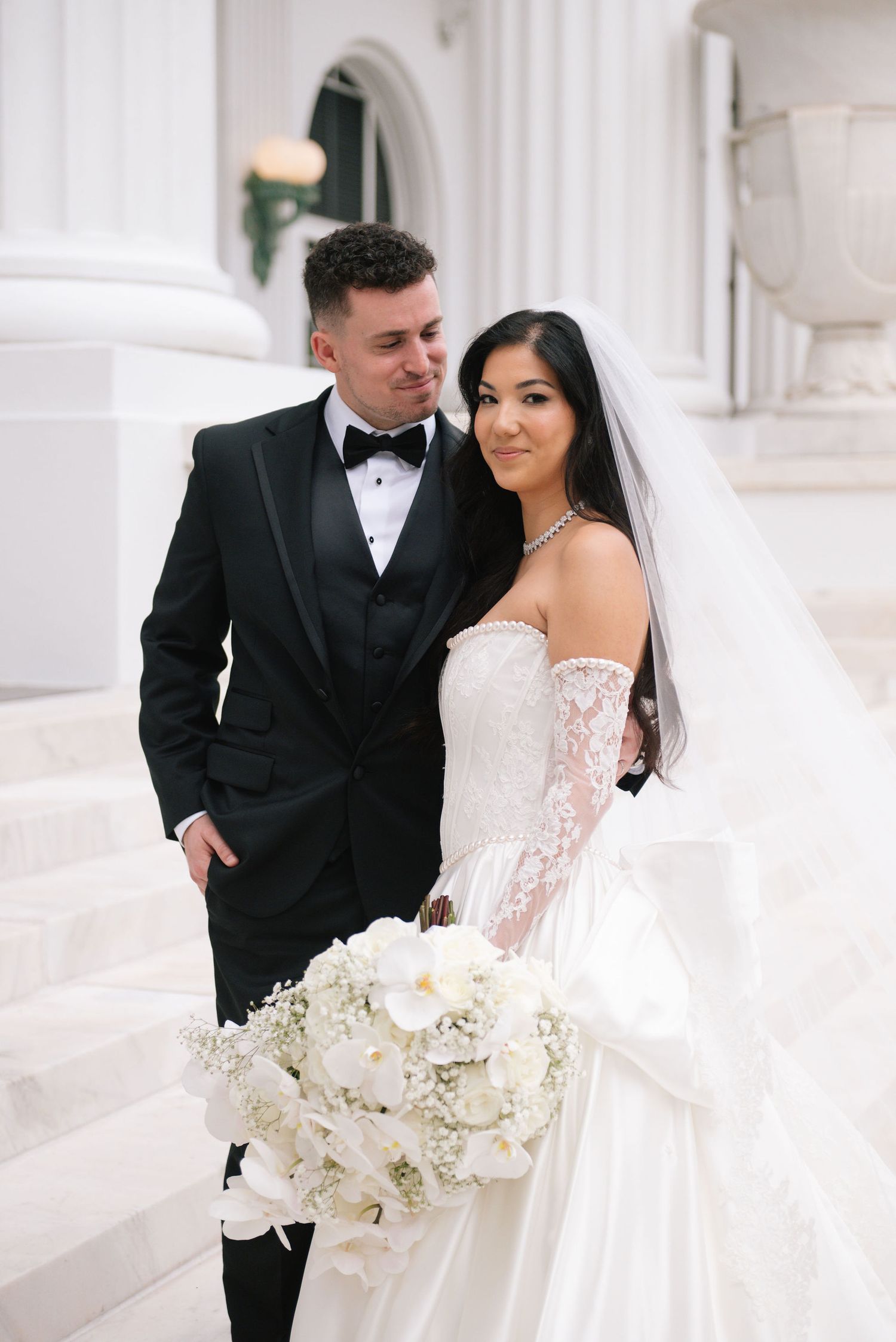 A bride holds a stunning white orchid bouquet while sharing a moment with her groom in front of white columns.