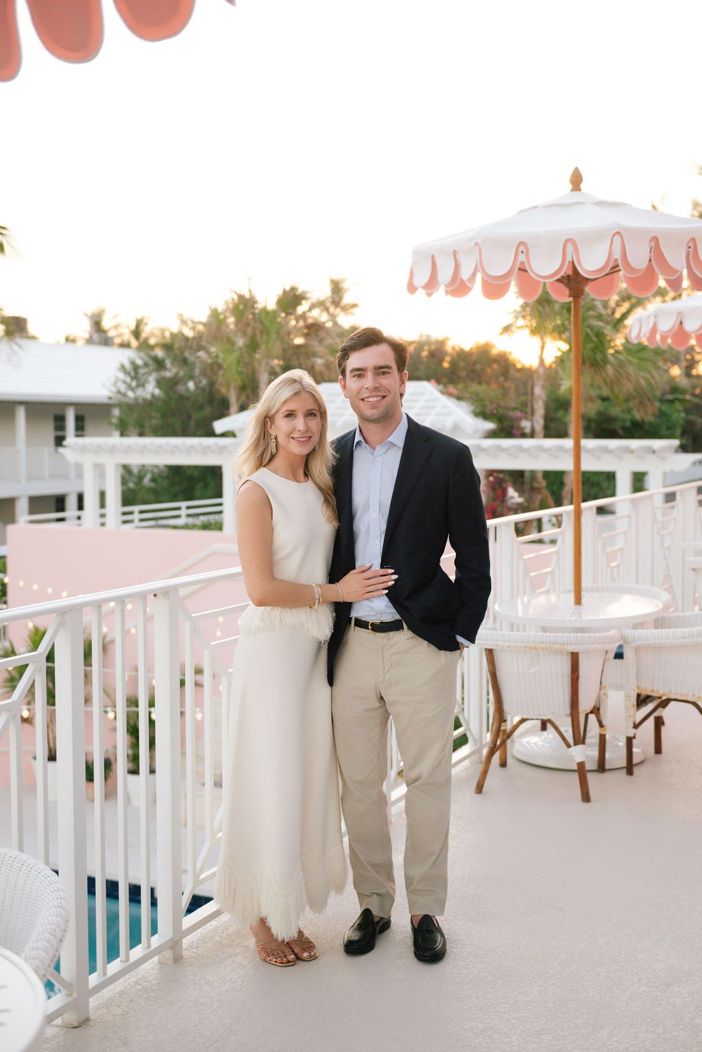 Elegant couple posing on balcony with decorative coral umbrella at sunset.