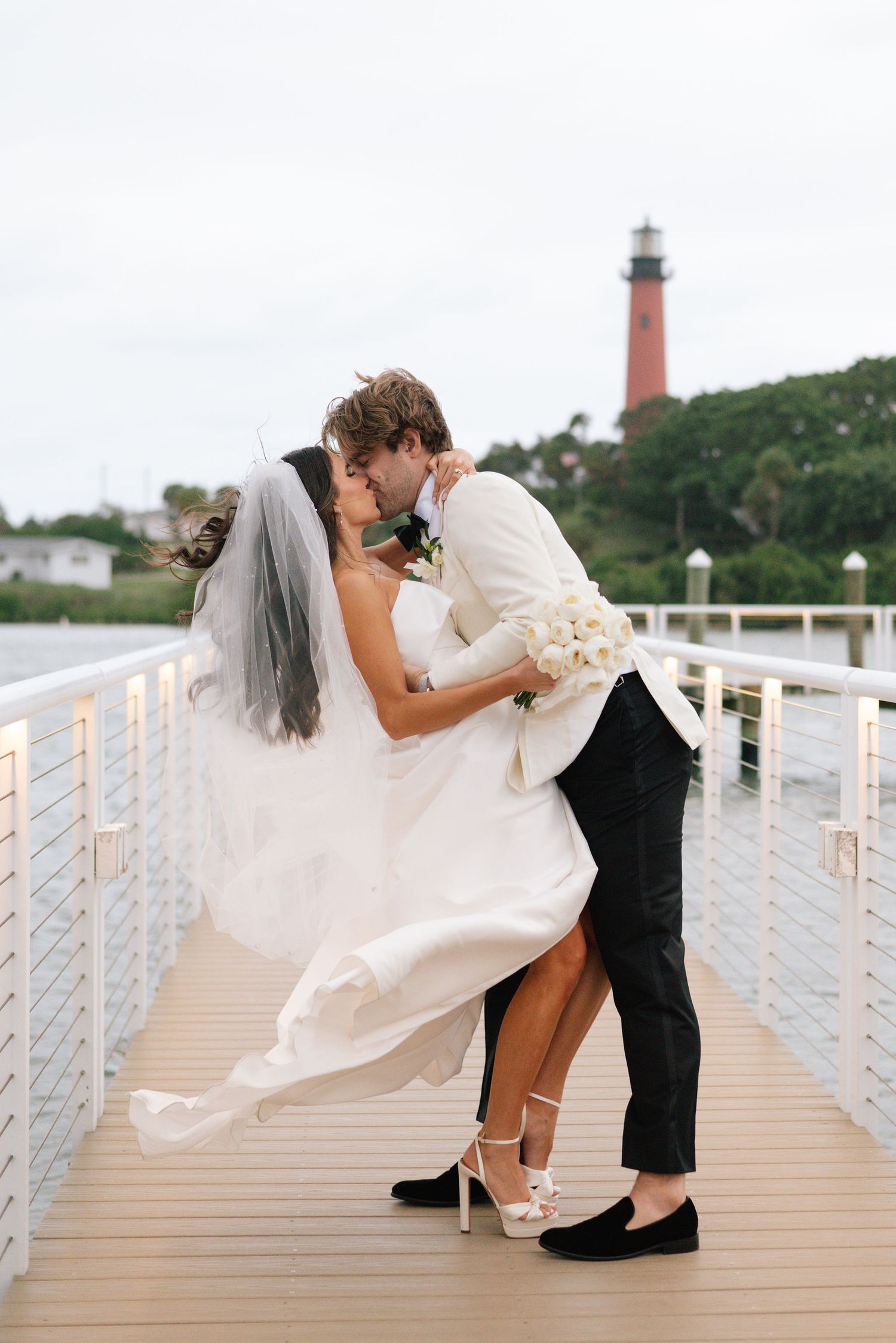 Wedding couple shares romantic moments on a waterfront boardwalk with lighthouse in background.