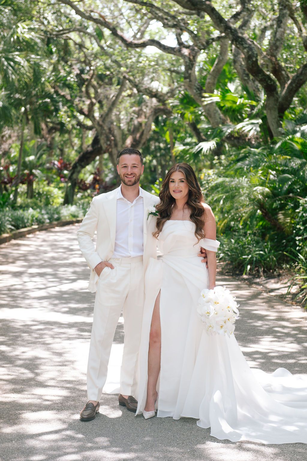 Wedding couple in all white attire walking along a tree-lined tropical path.
