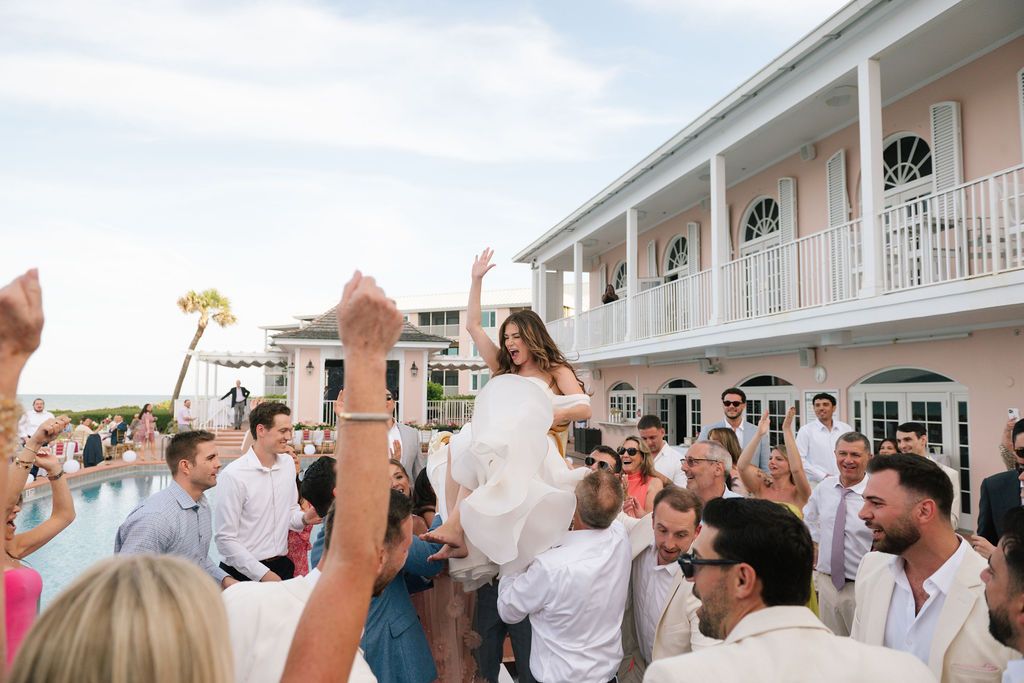 Wedding guests dance and celebrate on an outdoor patio at a beachside venue during sunset.