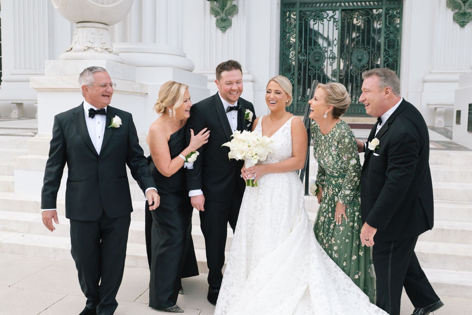 Wedding party in formal attire smiling and walking together outside elegant white building with green doors.