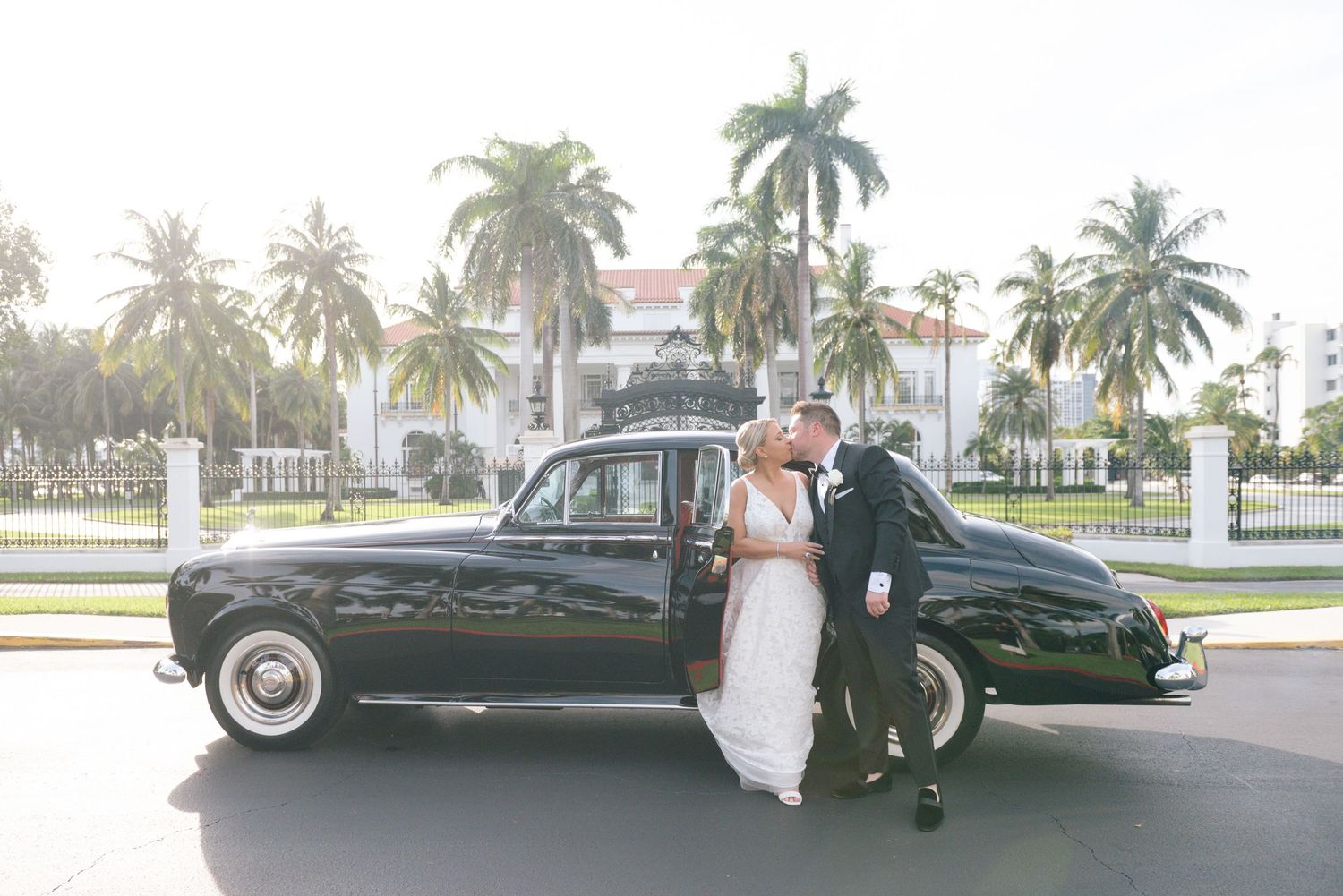 Bride and groom kissing beside a classic black vintage car with palm trees and elegant white mansion in background.