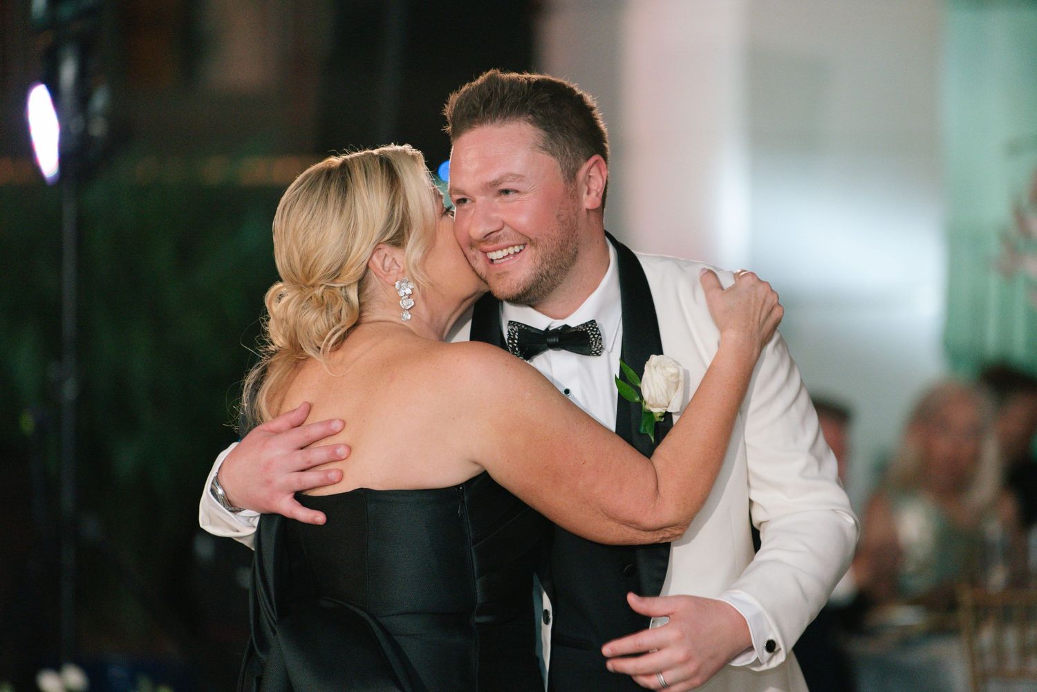 Groom in white jacket shares joyful embrace with woman in black dress at wedding reception.
