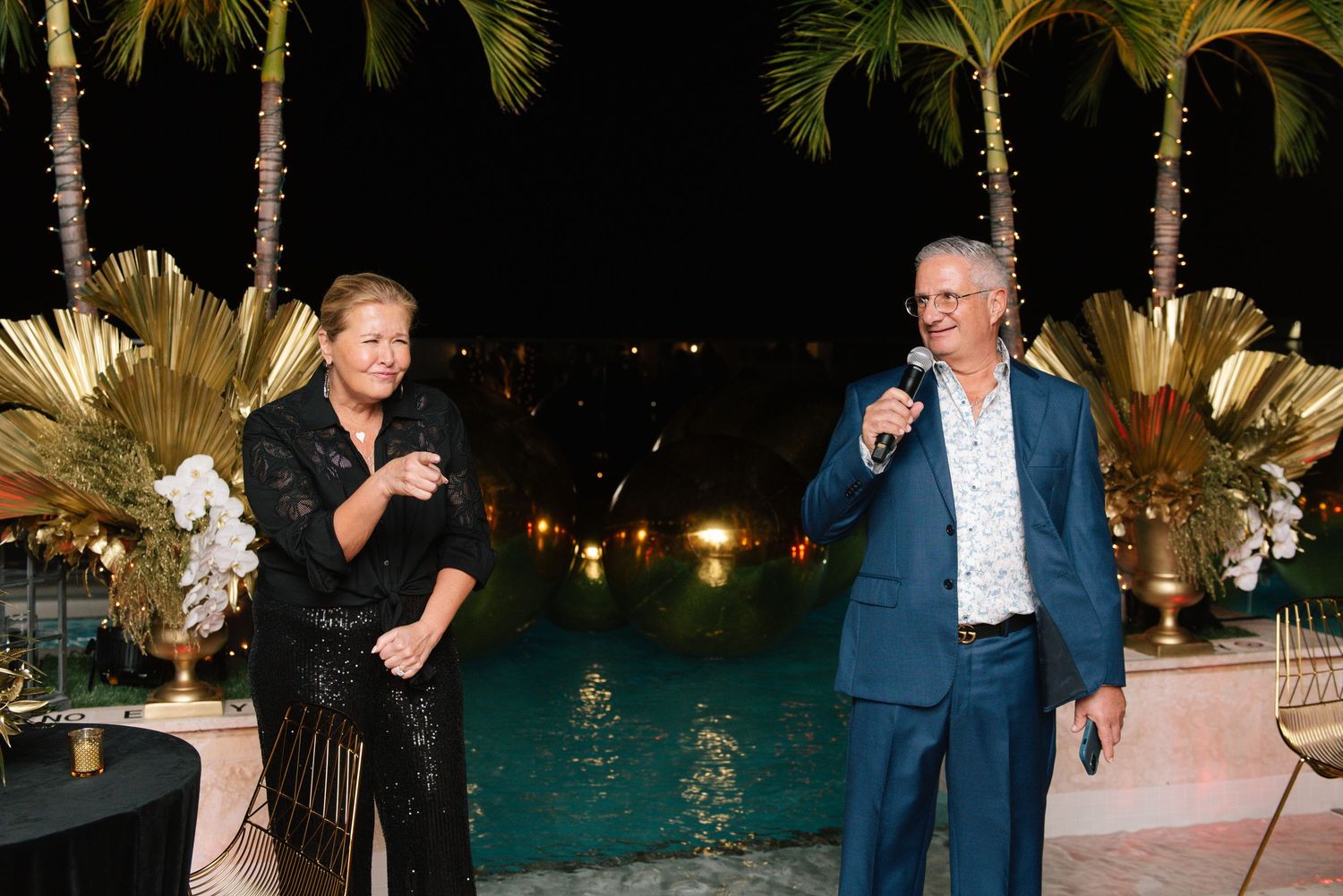 People gathered at an outdoor evening event near a pool with palm trees and decorative lighting.